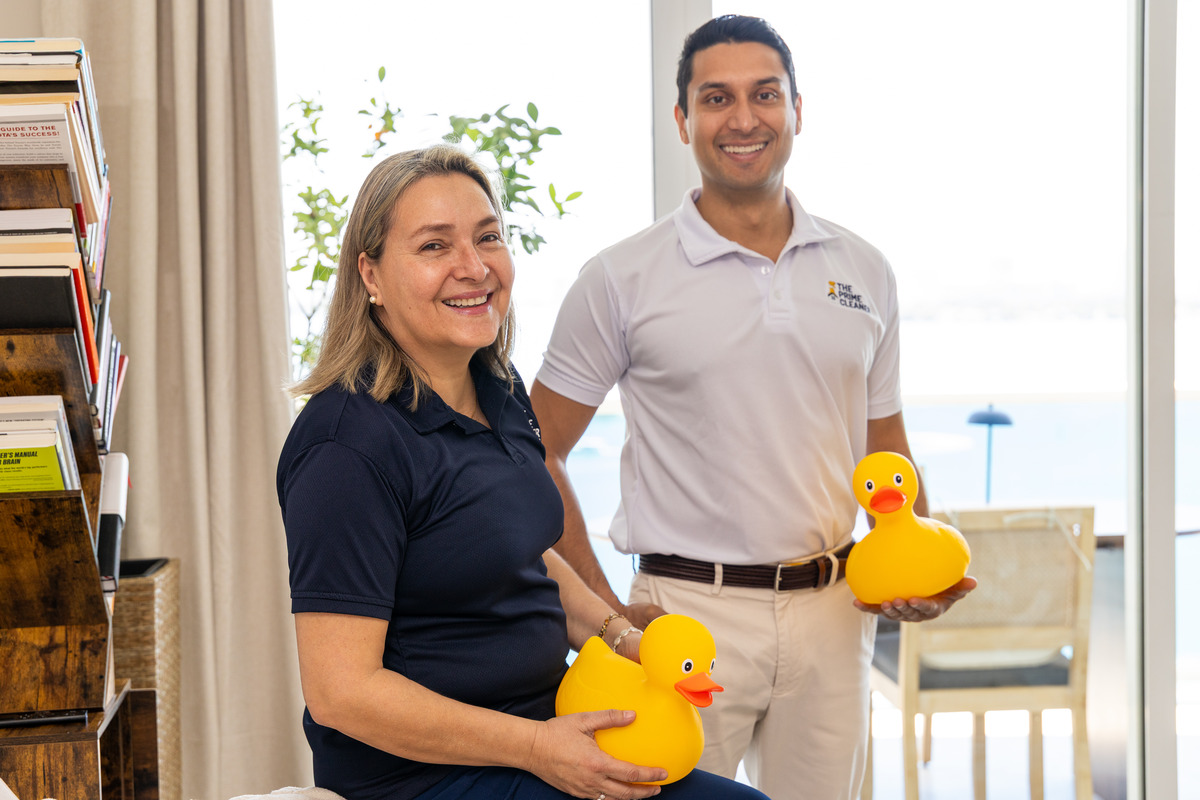 Two smiling people standing indoors near a window, holding yellow rubber ducks, with a bookshelf and a chair in the background.