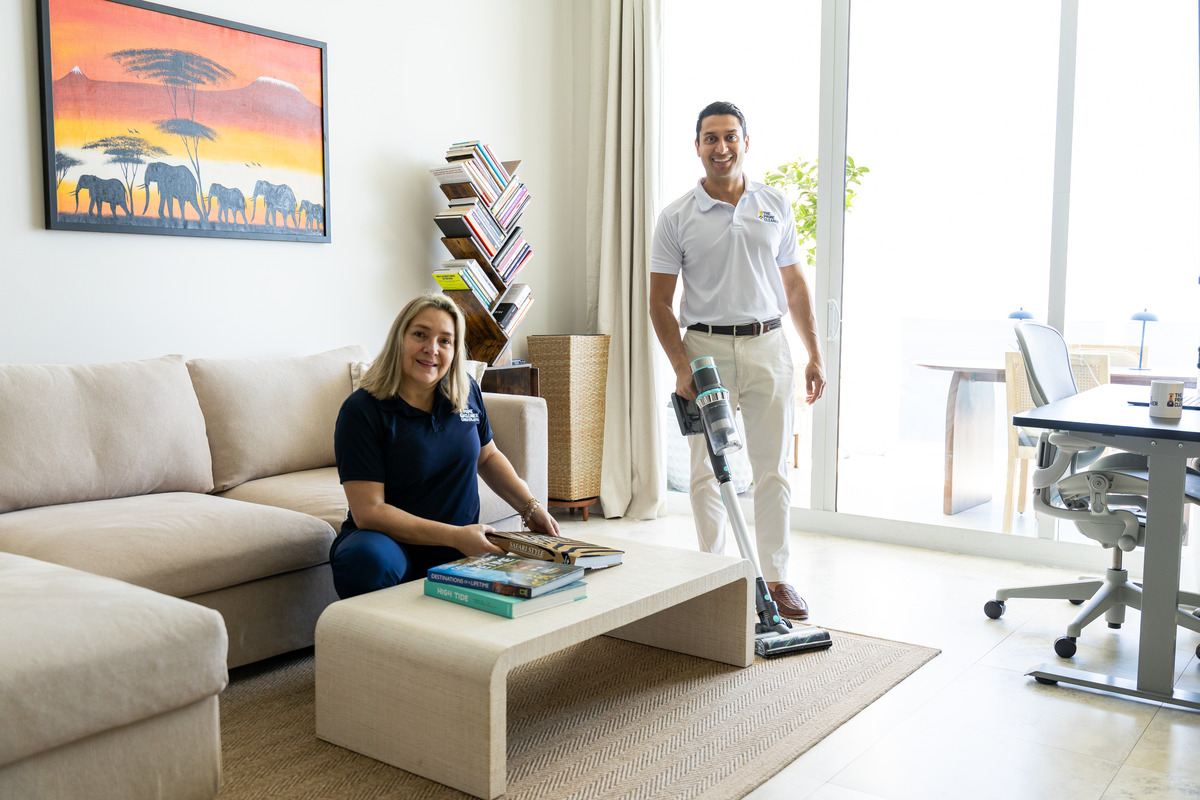 Two people in a modern living room, one sitting on a sofa with books, the other standing with a vacuum cleaner.
