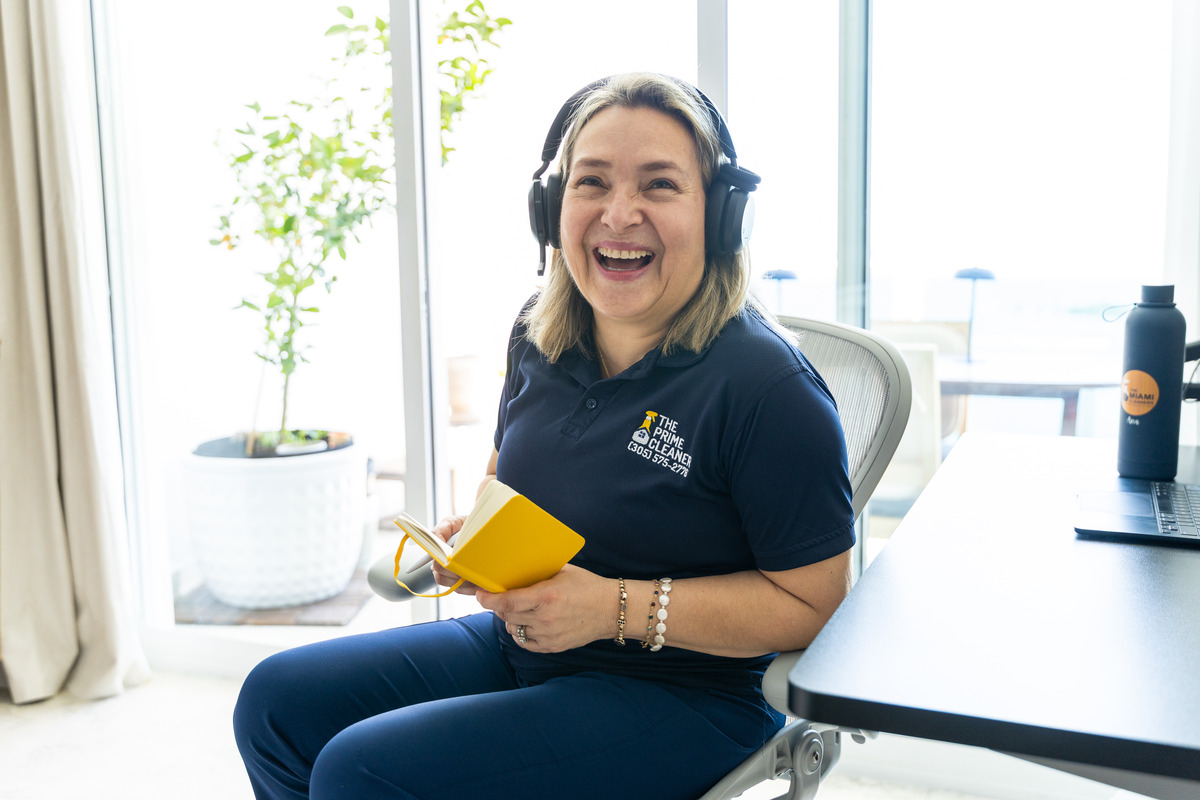 Woman smiling, wearing headphones, holding a yellow notebook, sitting at a desk in a bright room.