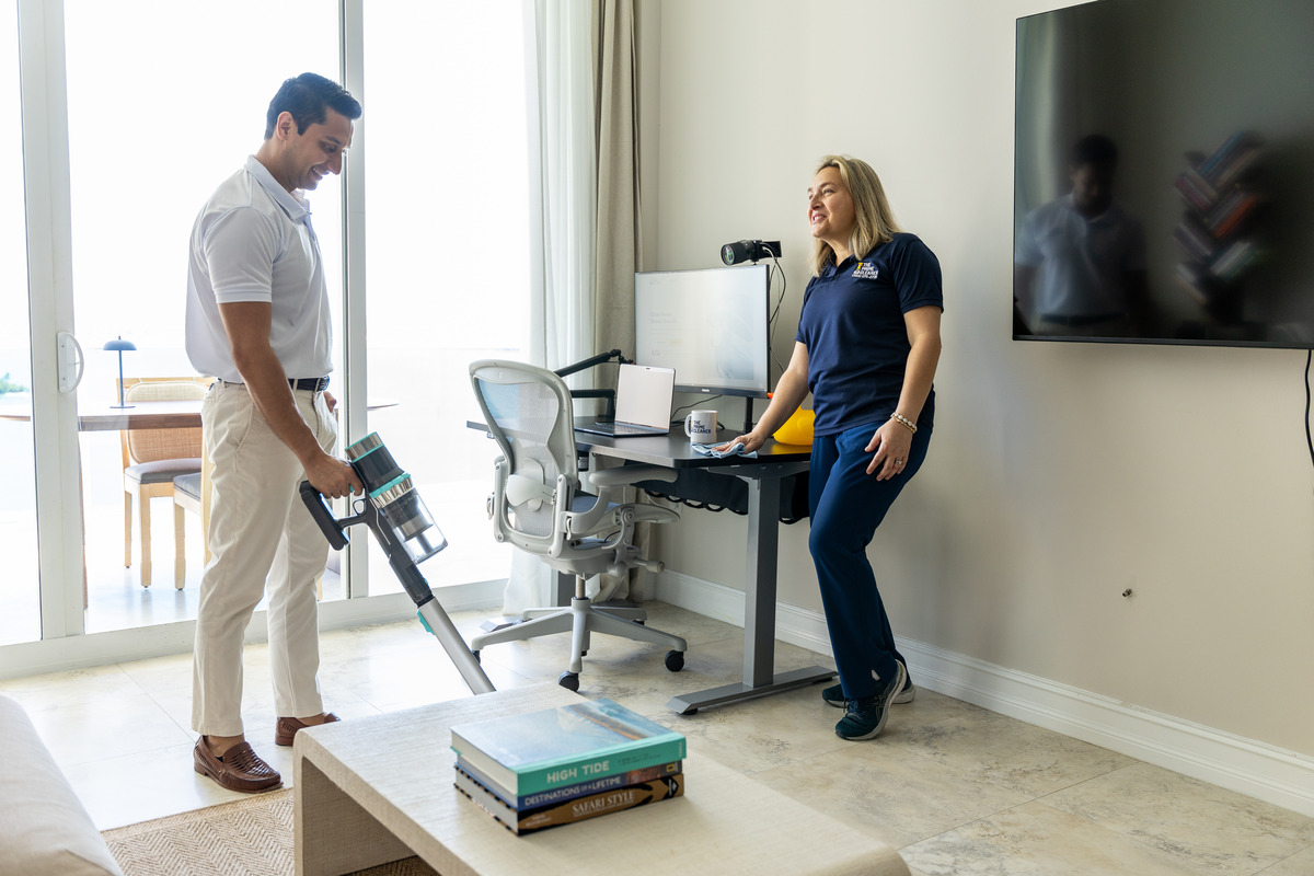 Two people in an office, one vacuuming the floor, the other standing near a desk and a large TV screen.