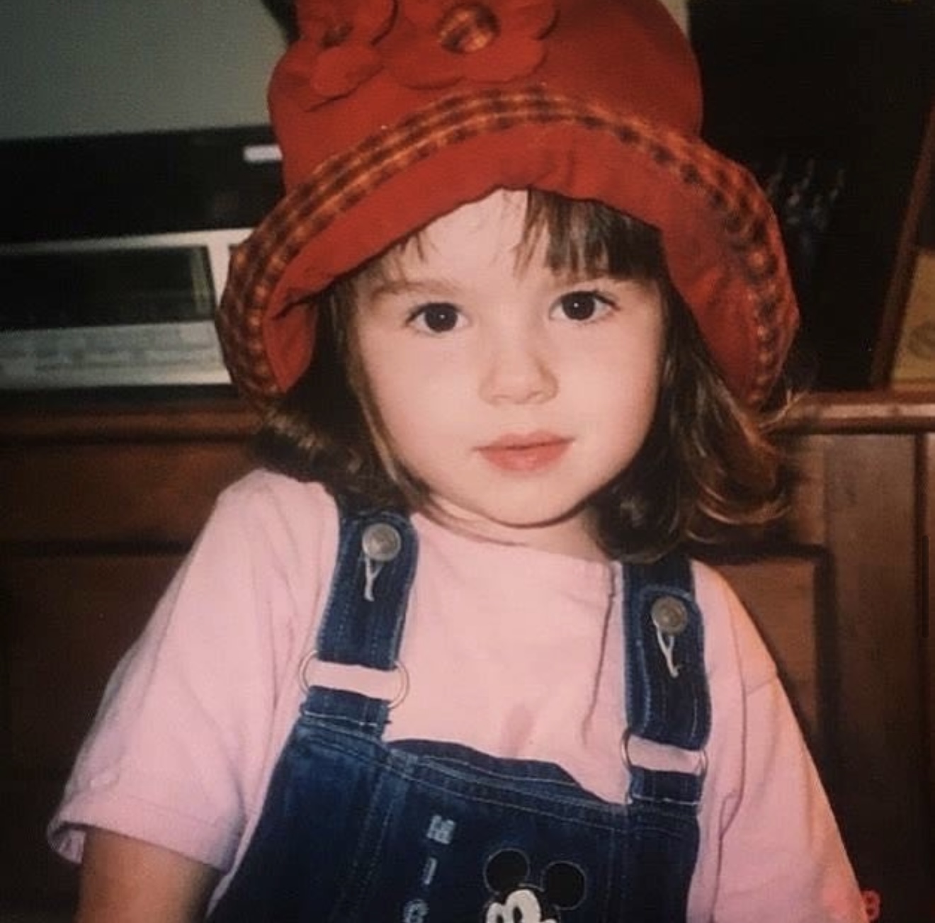 Young girl wearing a red hat, pink shirt, and denim overalls, looking at the camera.
