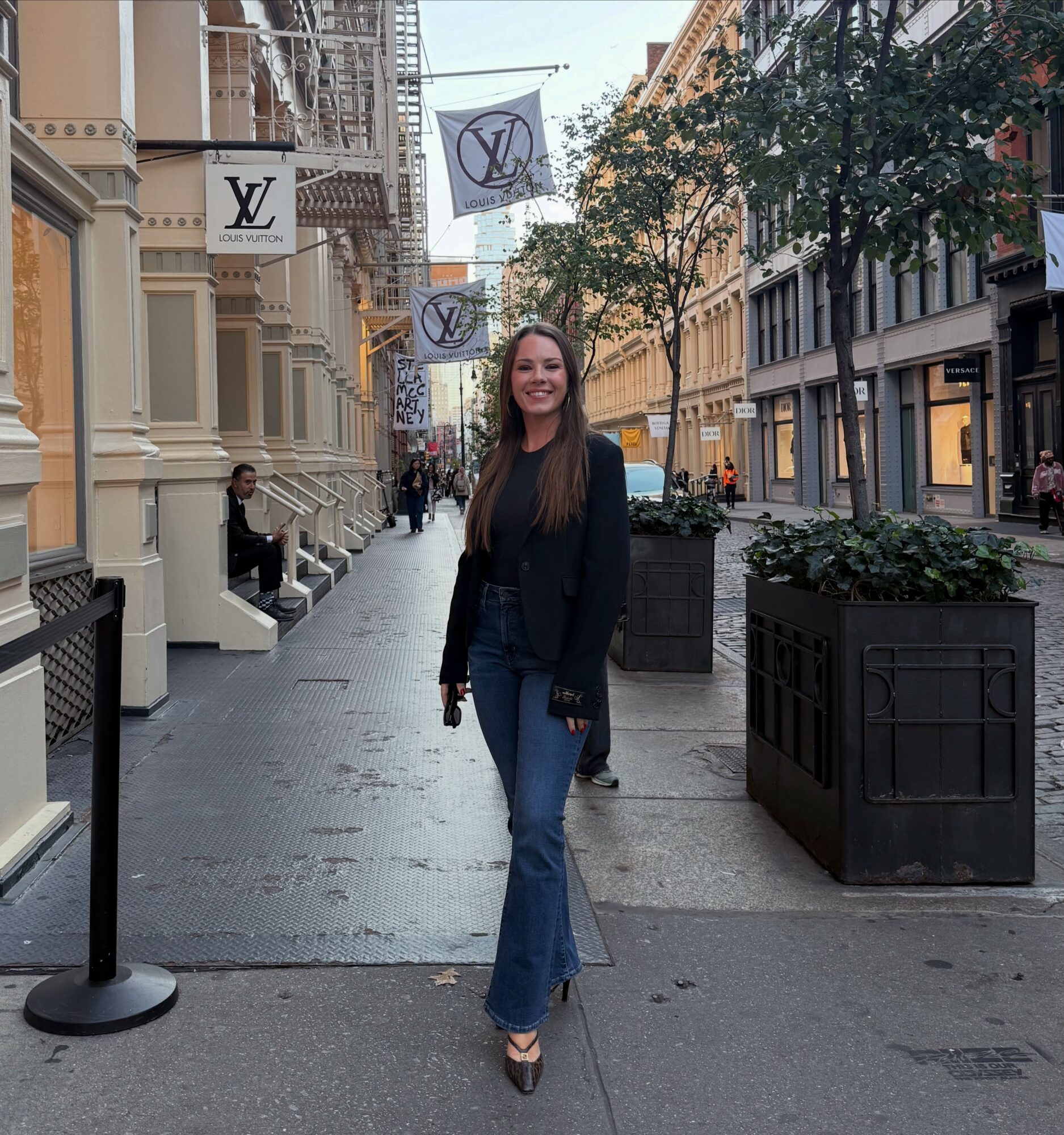 Young woman standing on city sidewalk, smiling, wearing jeans and a dark jacket, with buildings and trees in background.