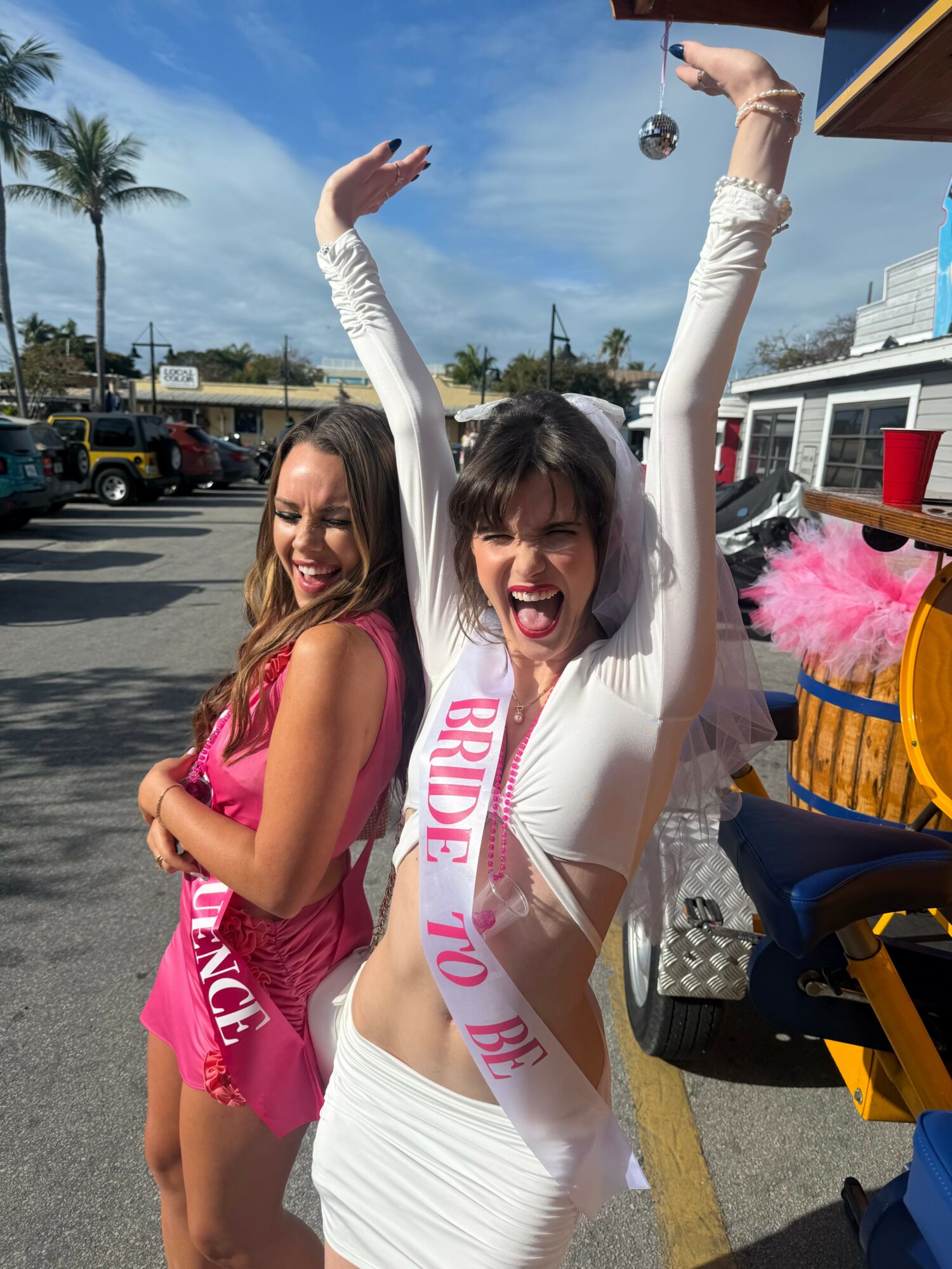 Two women celebrating outdoors, one wearing a sash that says 'Bride to Be,' smiling and raising arms.