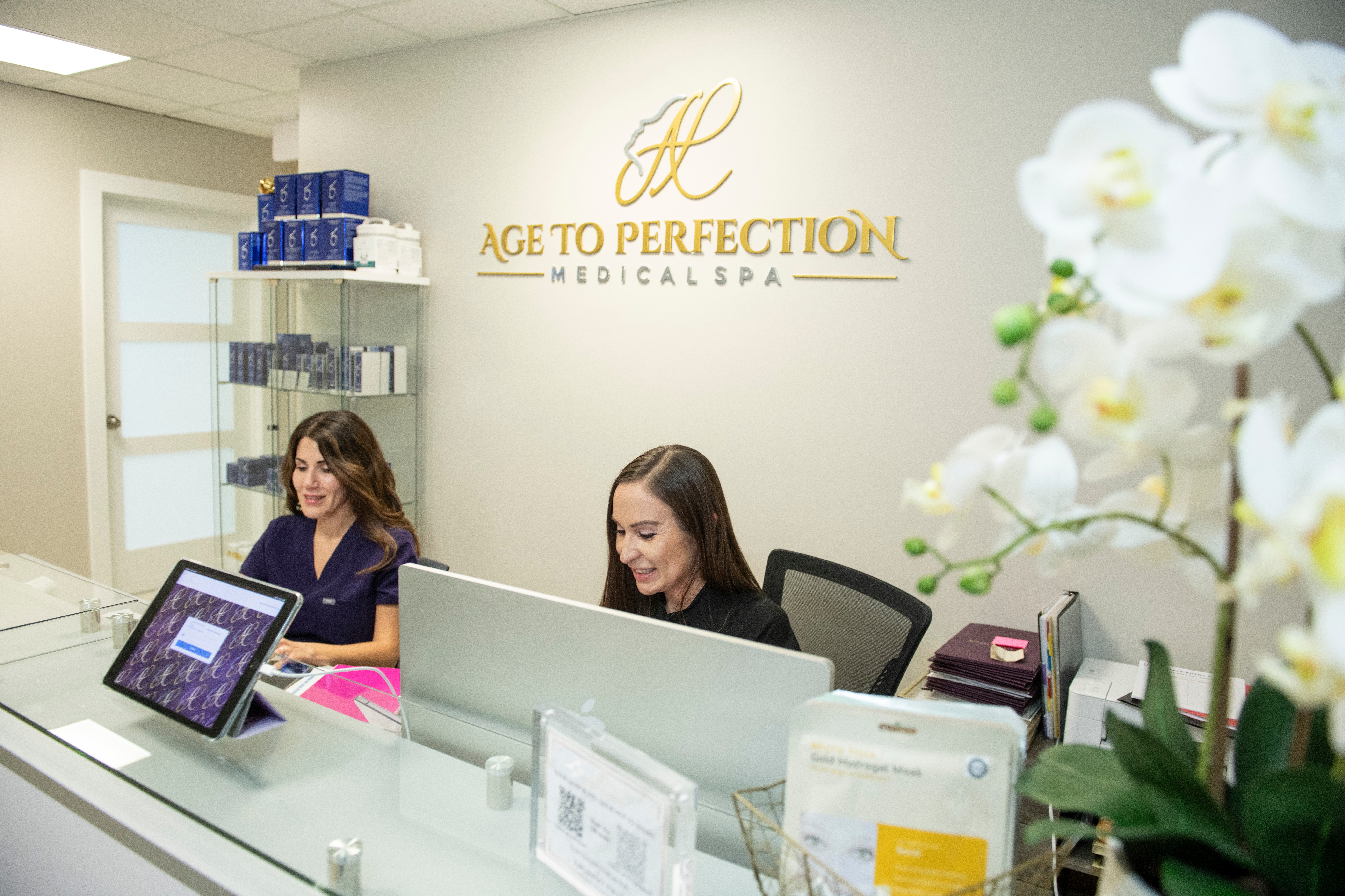 Two women sit at a reception desk in a medical spa, with a sign reading 'Age to Perfection Medical Spa' on the wall.