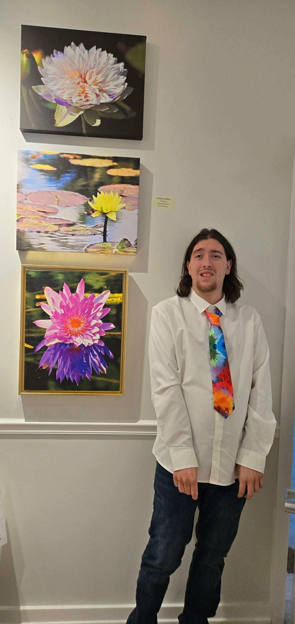 Person with long hair, white jacket, colorful tie, standing in front of three floral paintings on wall.
