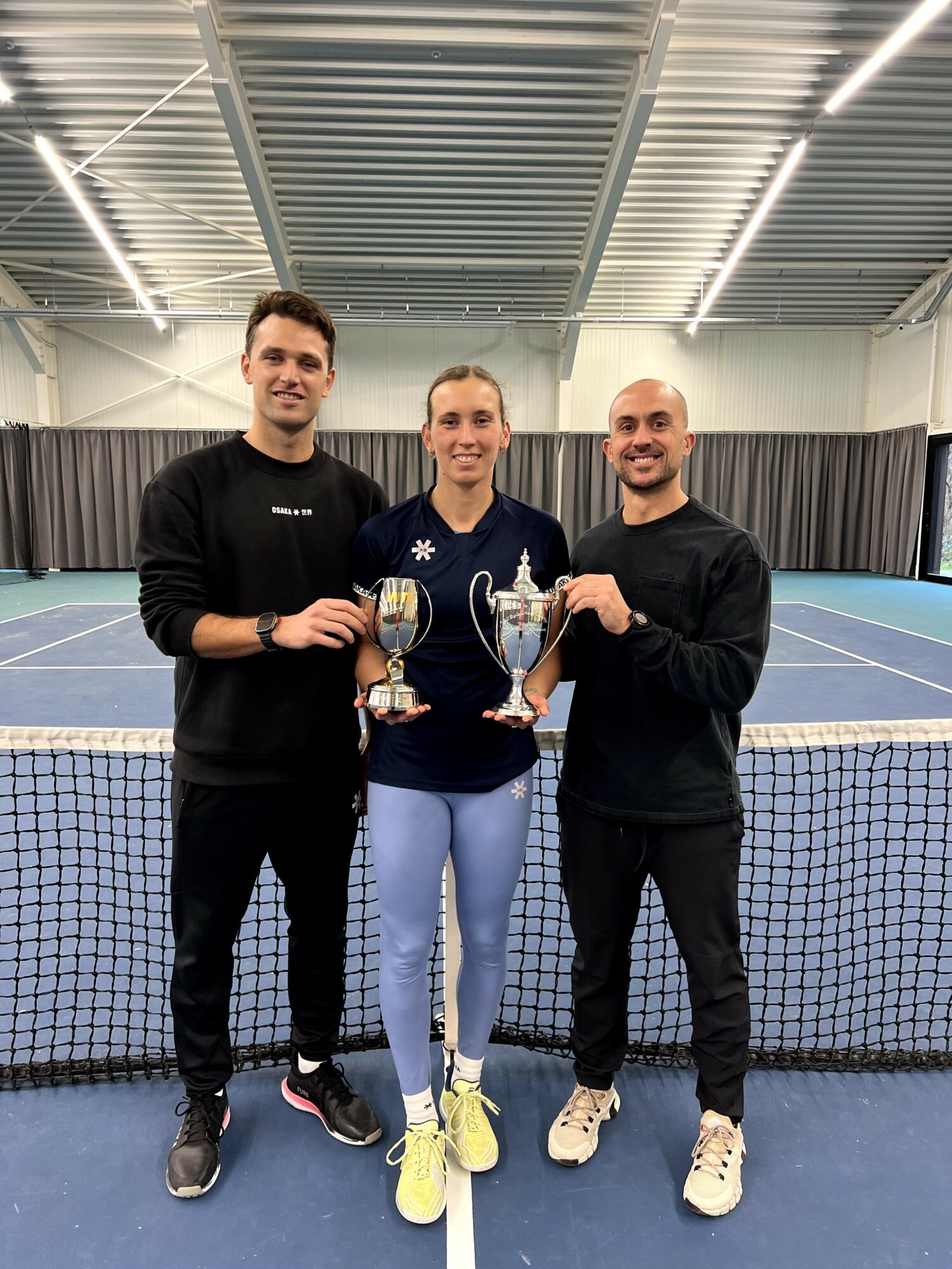 Three people holding trophies on an indoor tennis court, smiling, with a net in front and a curtain behind.