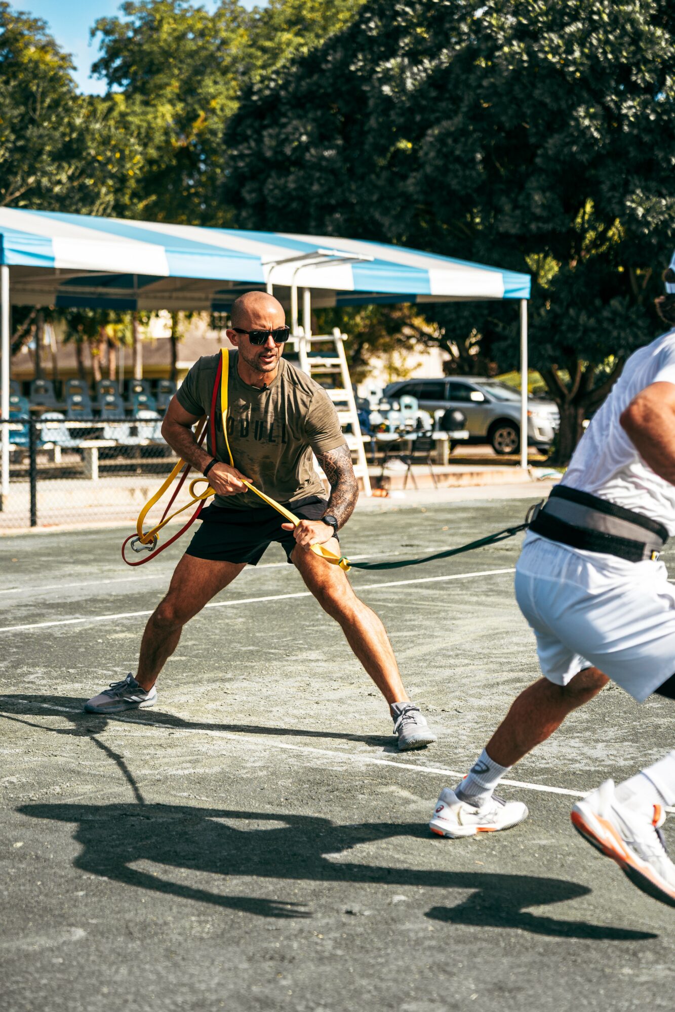 Person in sunglasses pulls a rope during a tug-of-war game outdoors, with trees and parked cars in background.