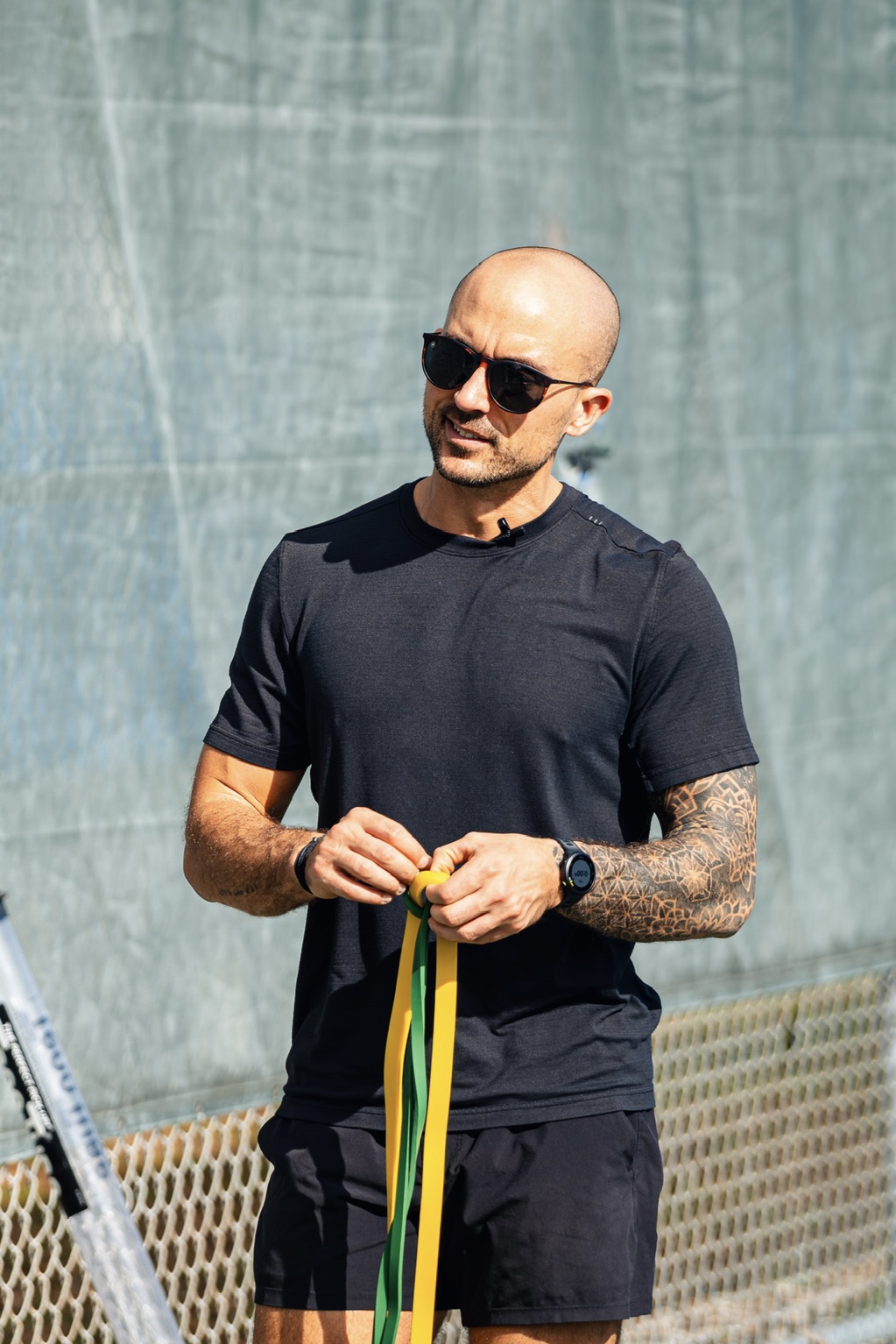 Bald man wearing sunglasses, black t-shirt, and shorts, holding a yellow and green ribbon outdoors.