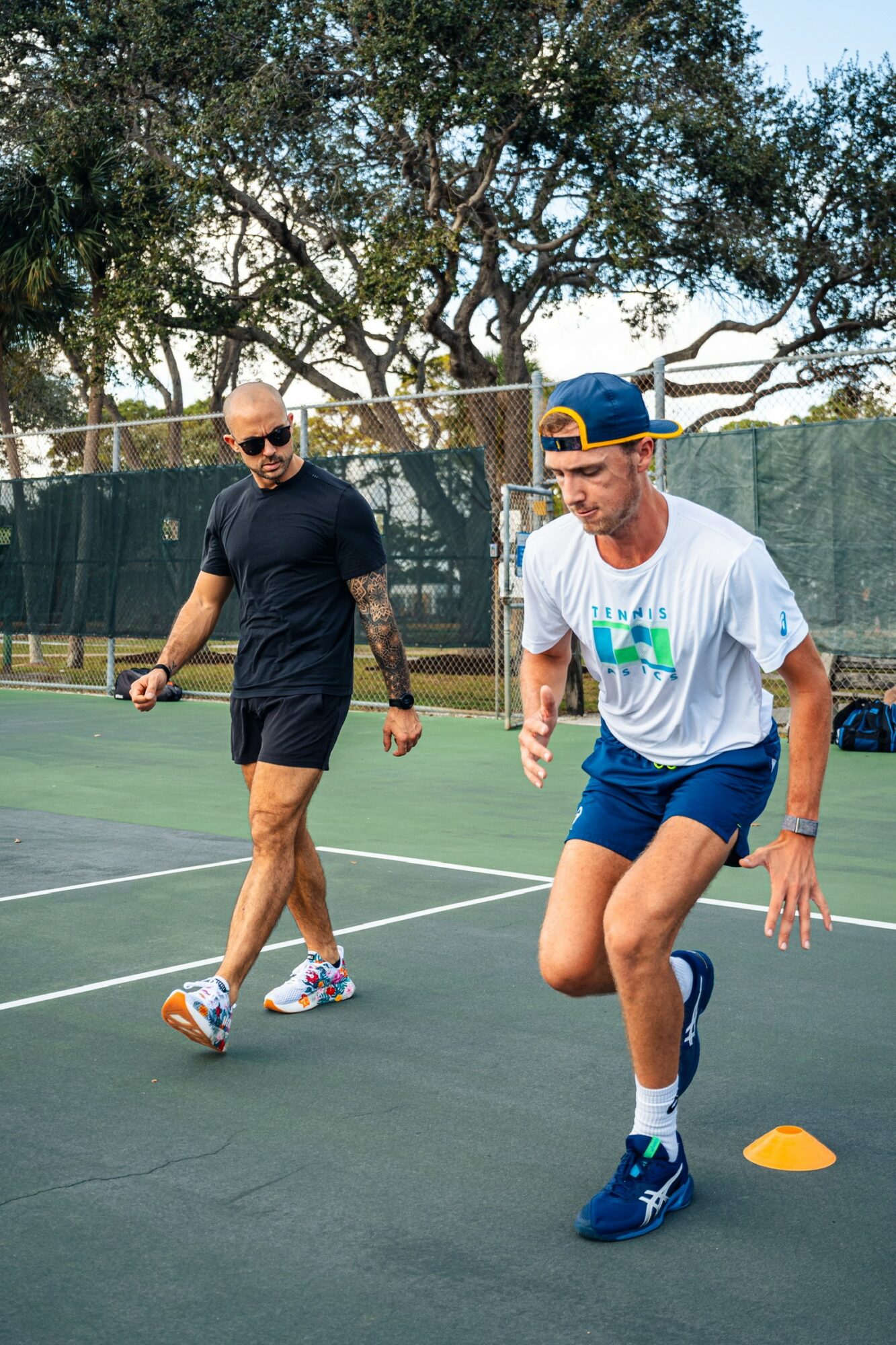 Two men on a tennis court, one running around a cone, the other walking nearby, trees in background.