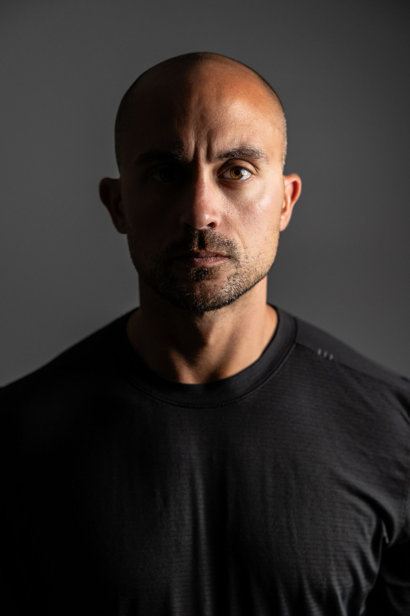 Man with a serious expression, bald head, and wearing a black shirt, facing forward against a dark background.