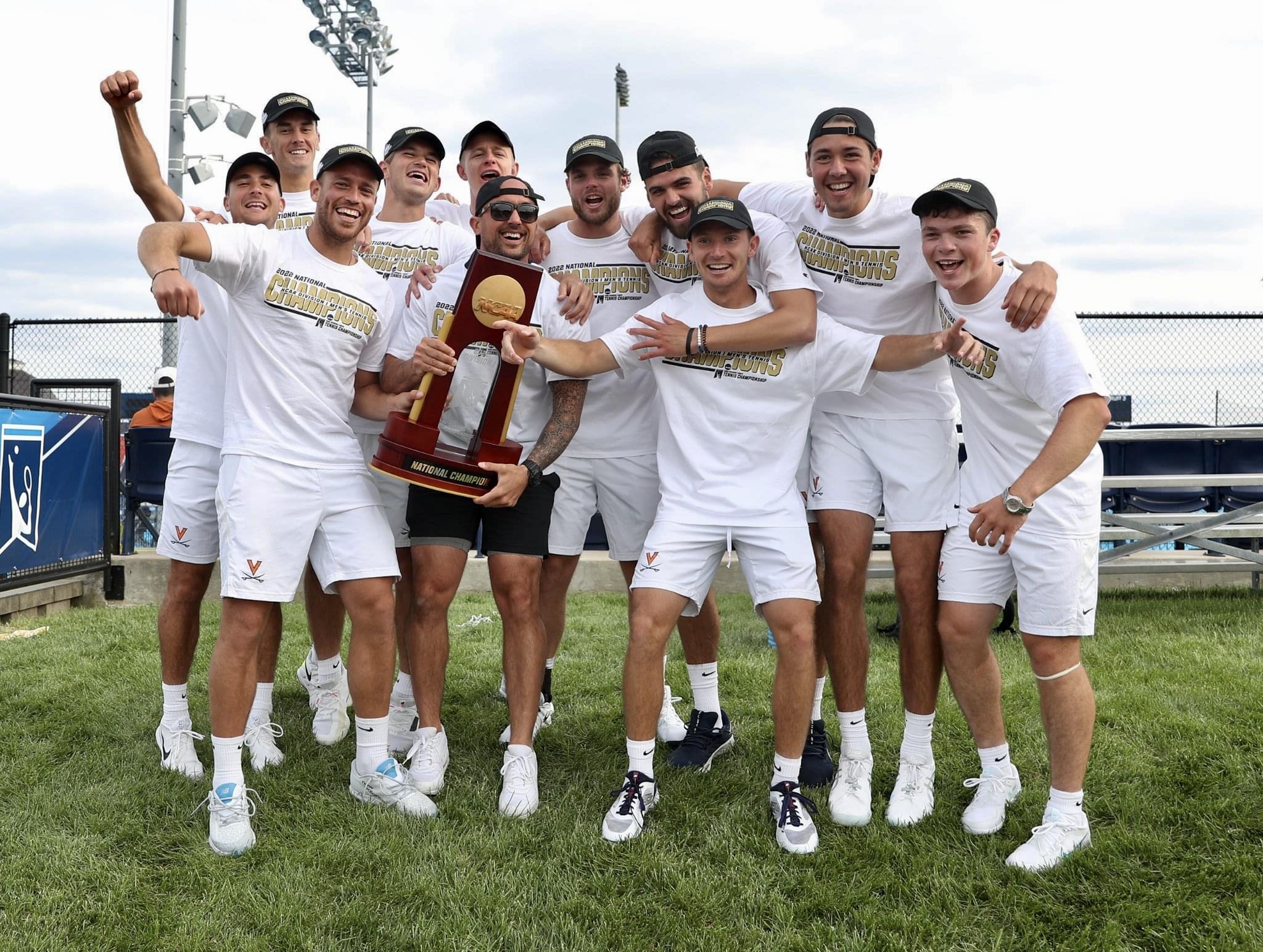 Group of ten people in white sportswear holding a trophy on a grassy field, celebrating.