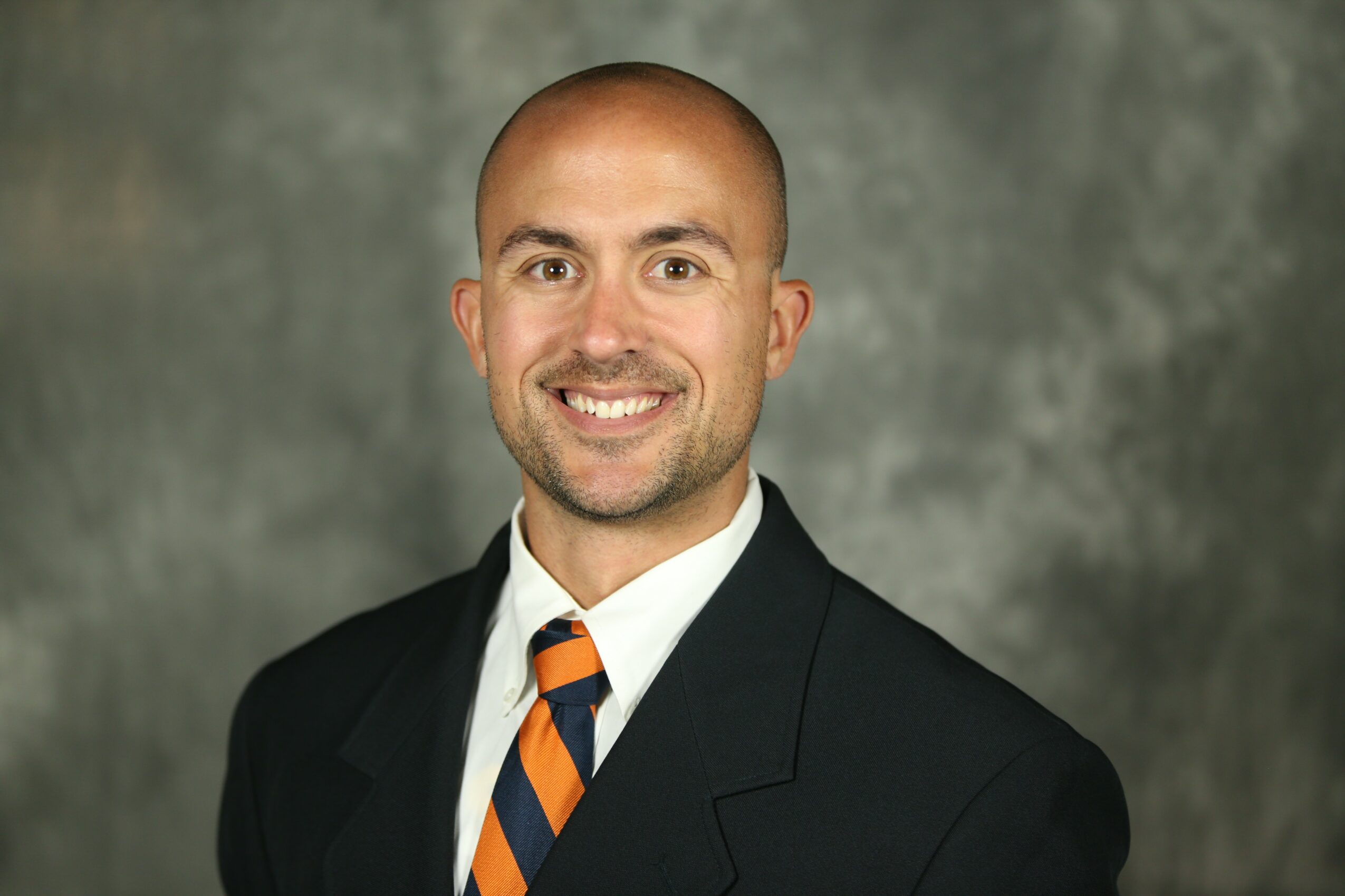 Man with a shaved head and beard wearing a suit and tie, smiling against a gray background.