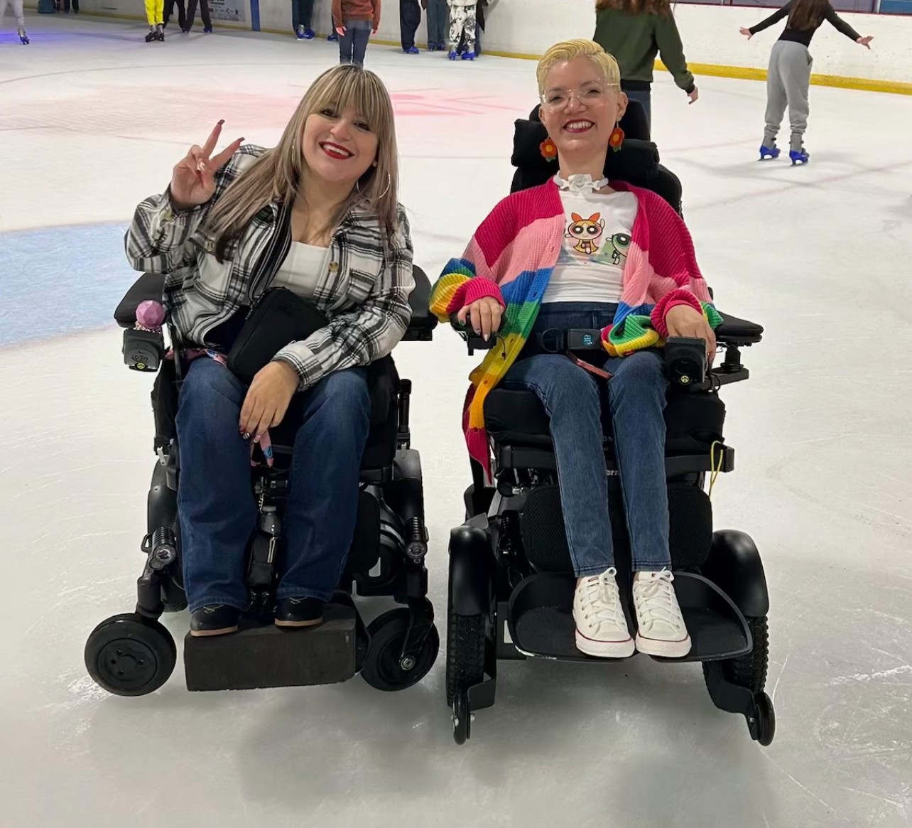 Two women in wheelchairs on ice rink, smiling, one making peace sign, background with people skating.