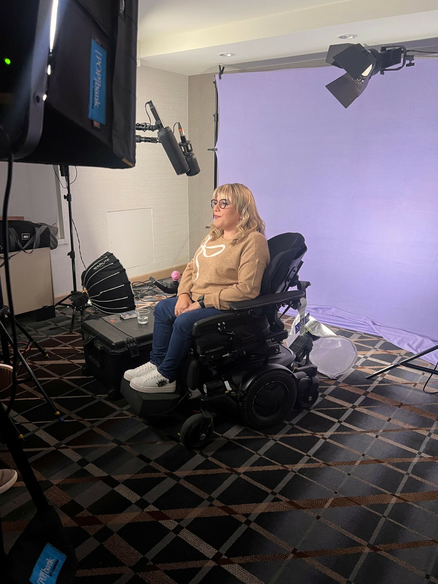 Woman in beige sweater and glasses seated in a motorized wheelchair in a studio with a purple backdrop and professional lighting.