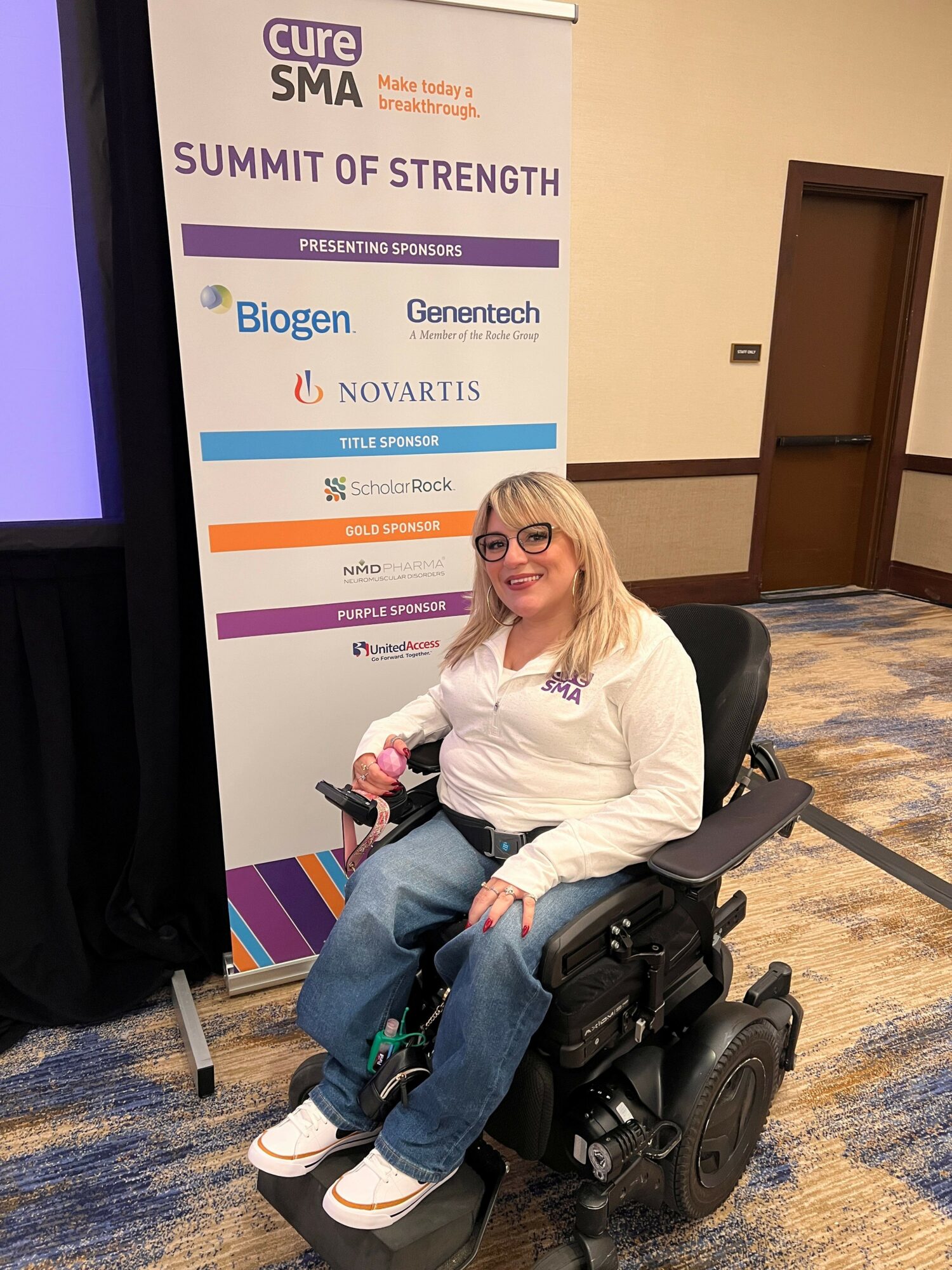 Woman in a wheelchair smiling, wearing glasses and a white shirt, seated in front of a conference banner.