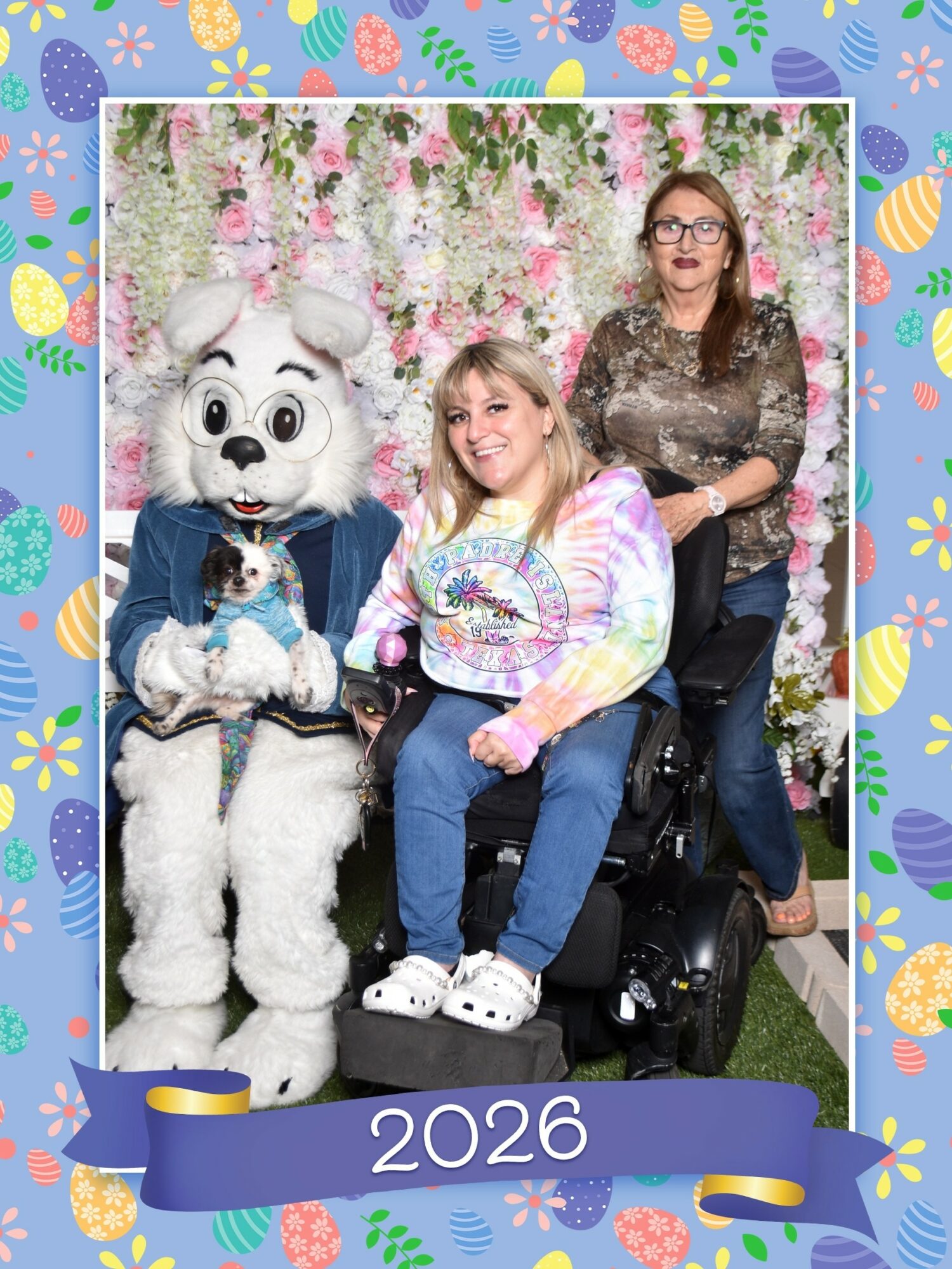 Two women and a person in a dog costume with a small dog, in front of a floral backdrop, celebrating 2026.