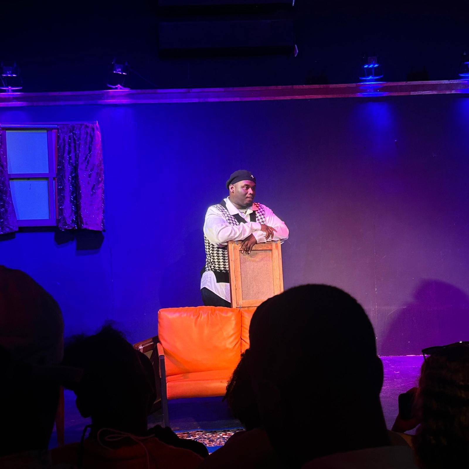 Performer standing behind a wooden podium on stage with an orange chair, audience silhouettes visible in foreground.
