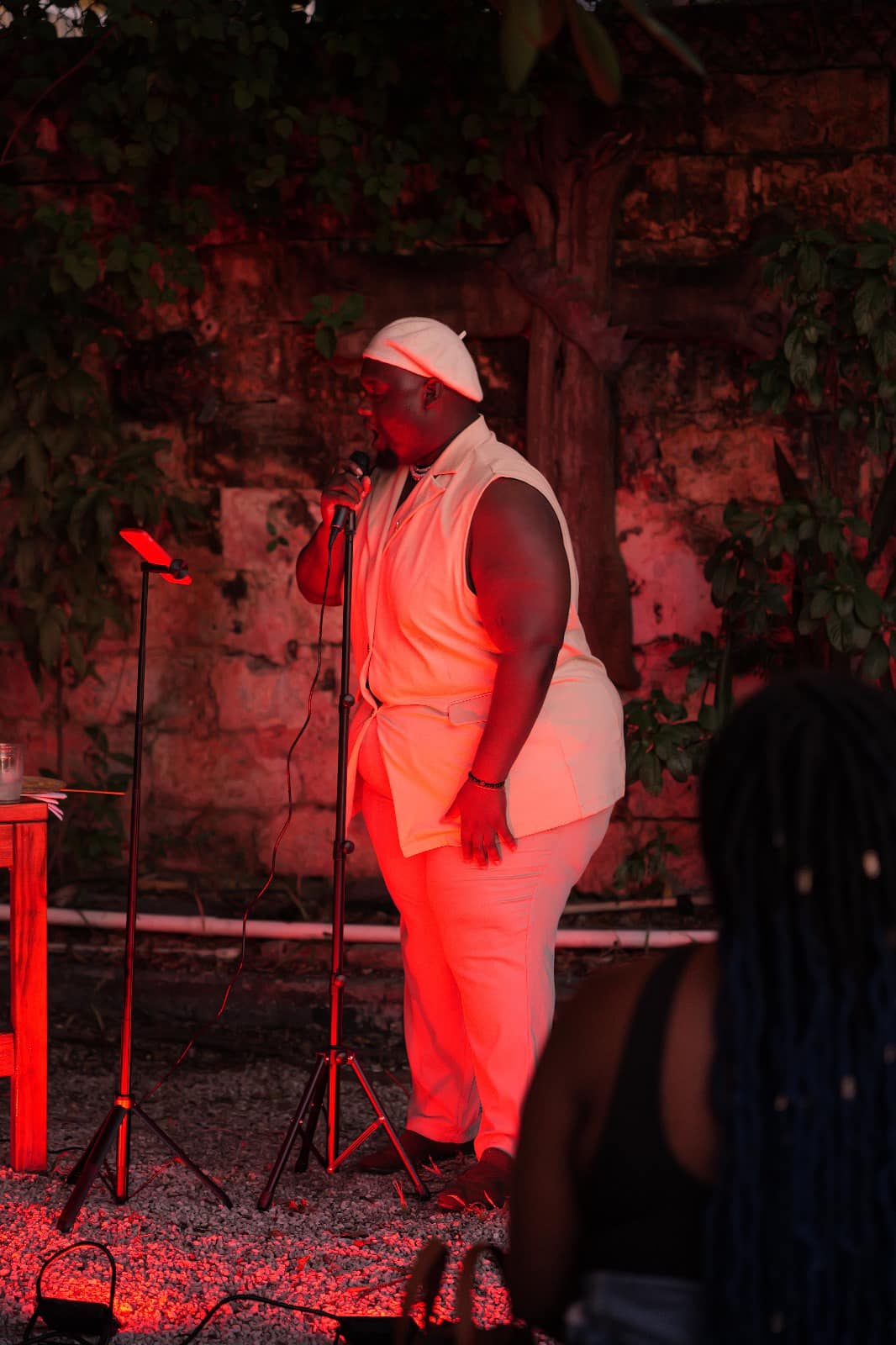 Person wearing white outfit and cap standing outdoors at night with microphone and equipment nearby.