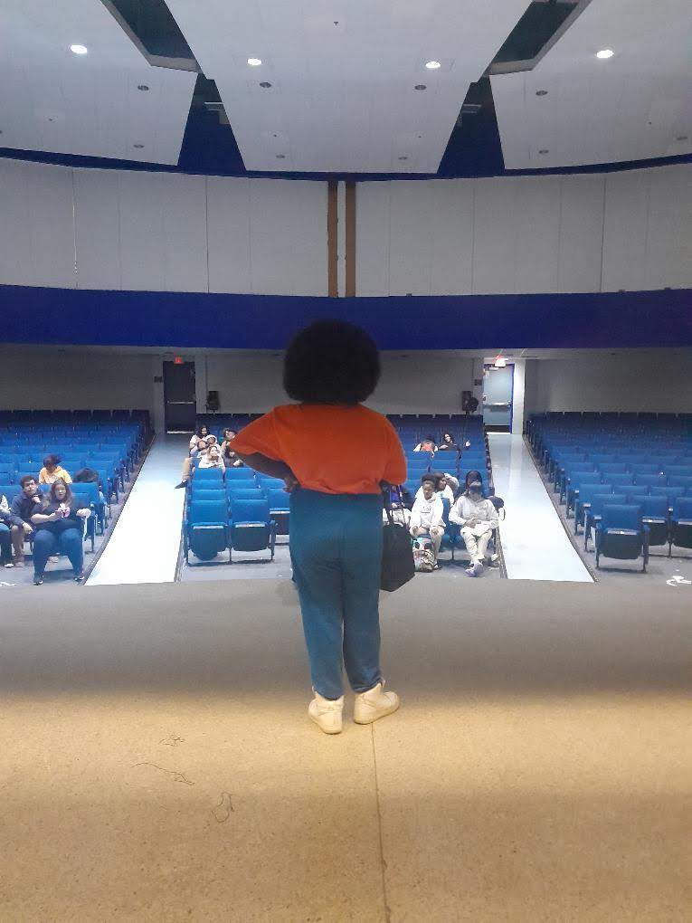 Person with curly hair, orange top, and blue jeans standing on stage facing an audience in a large auditorium.