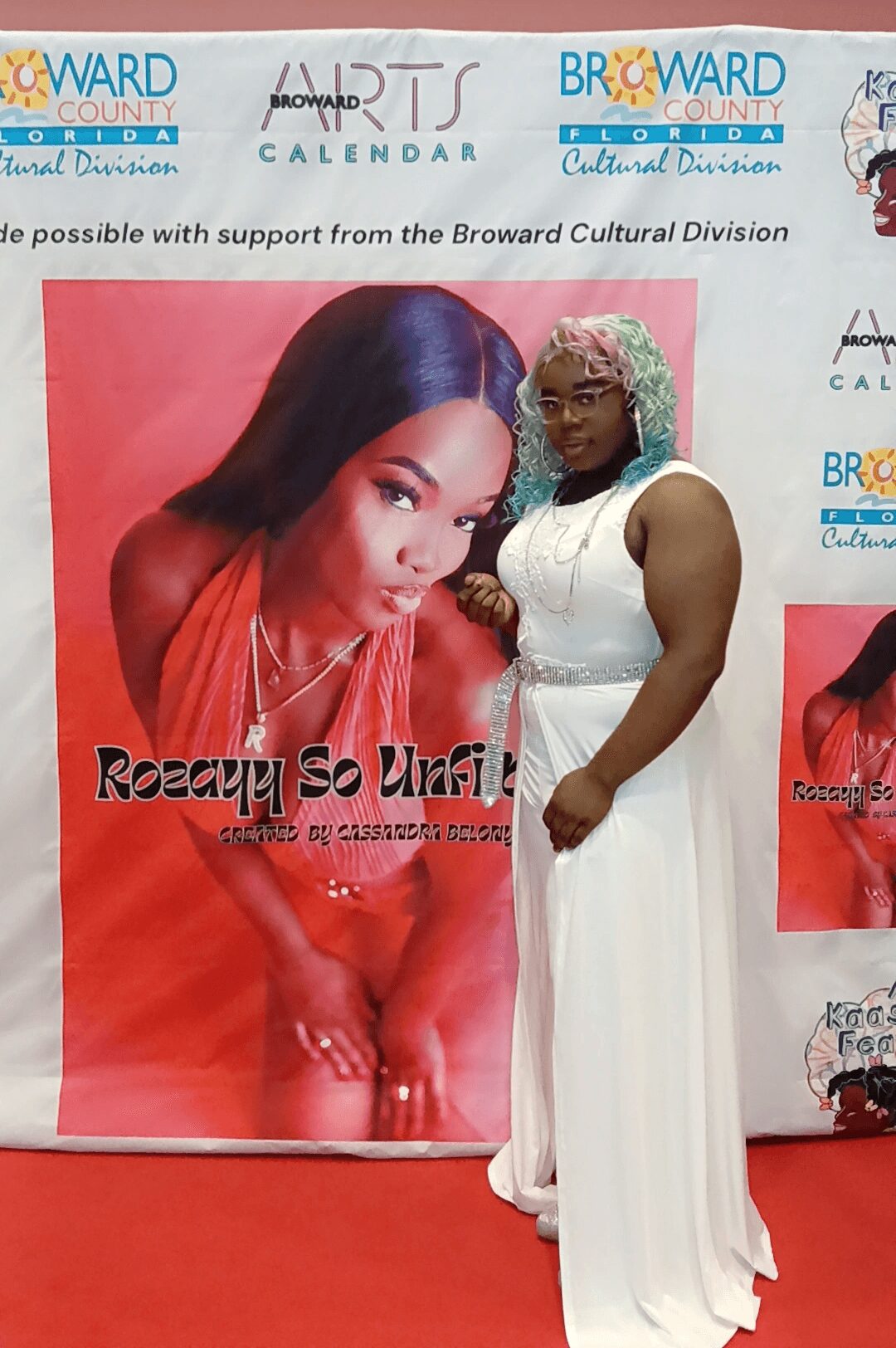 Woman in white dress standing next to a large poster of a woman with long dark hair, red background.