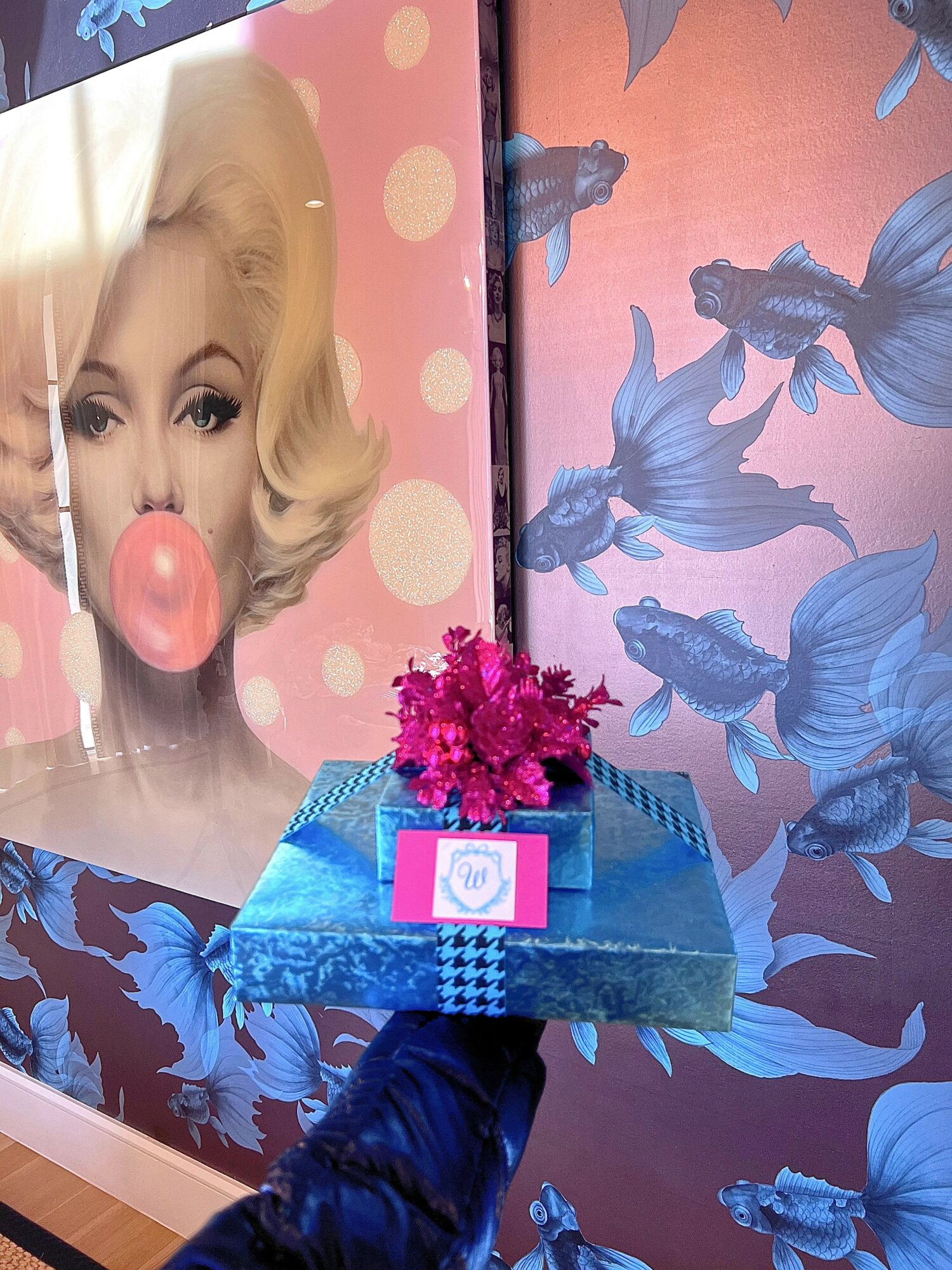 Person holding a tray with pink flowers in front of a Marilyn Monroe poster and blue fish-patterned background.