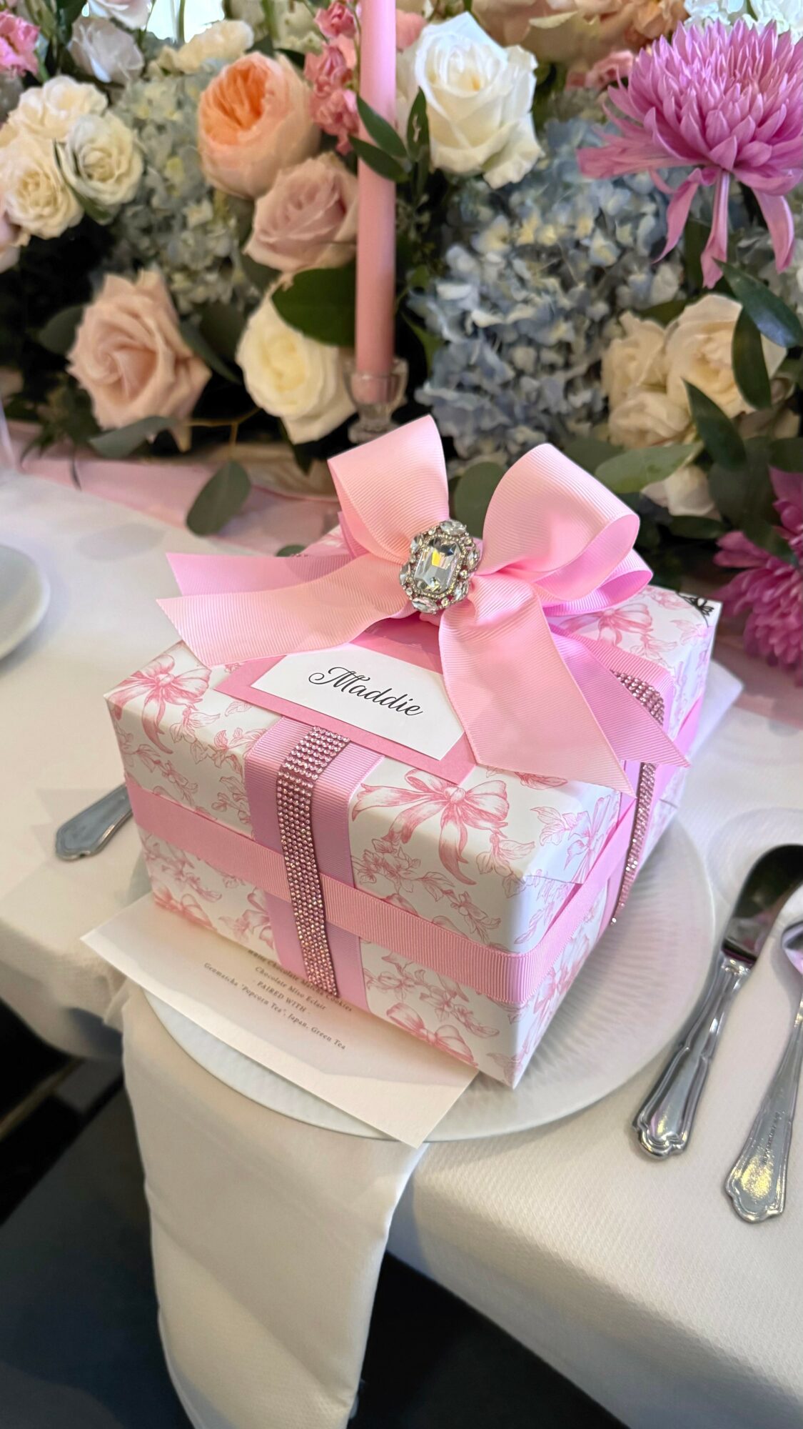 Decorative gift box with pink ribbon and bow, surrounded by flowers on a table.