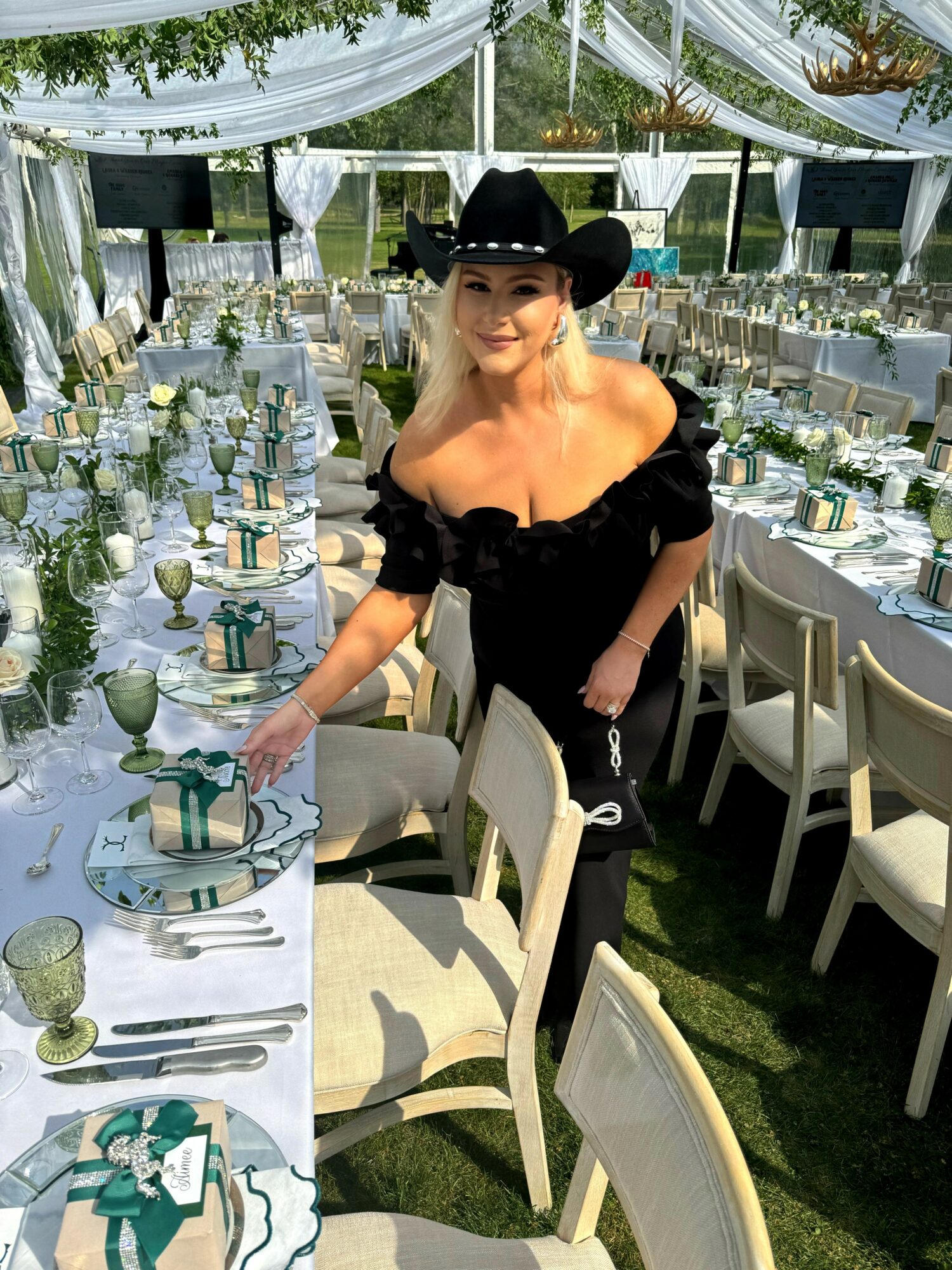Woman in black dress and cowboy hat arranging table settings outdoors under a tent.