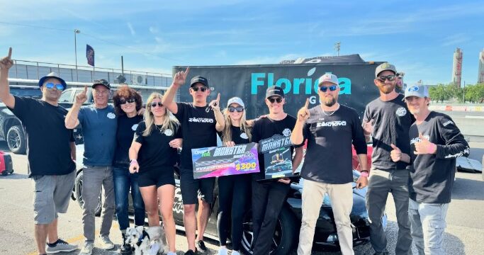 Group of people standing outdoors in front of a bus, holding signs, some wearing sunglasses and casual clothing.
