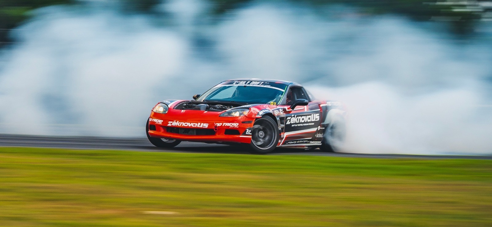 Race car drifting on track with smoke behind, grass in foreground, blurred background.