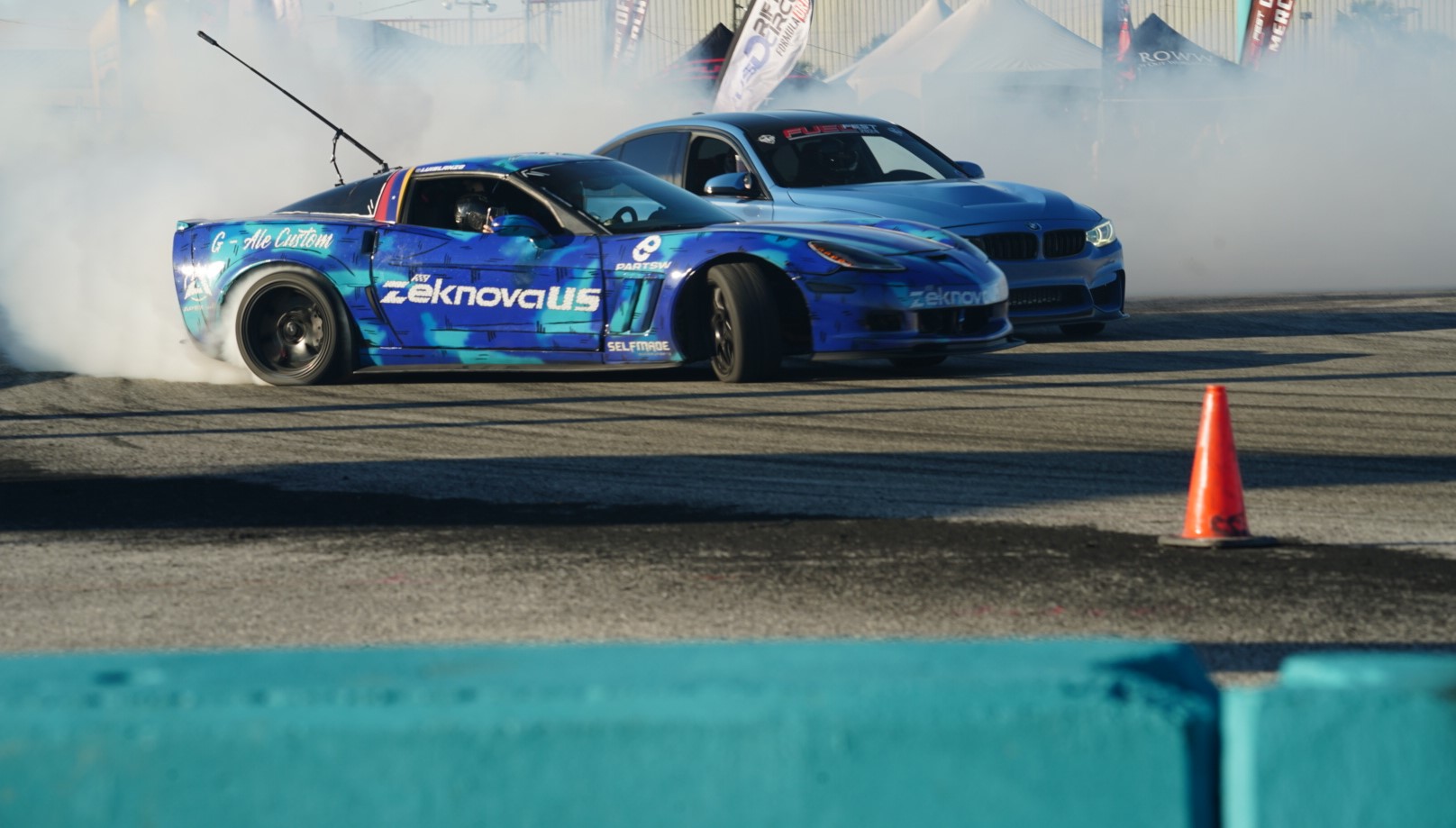 Two cars racing on a track with smoke and an orange cone nearby.