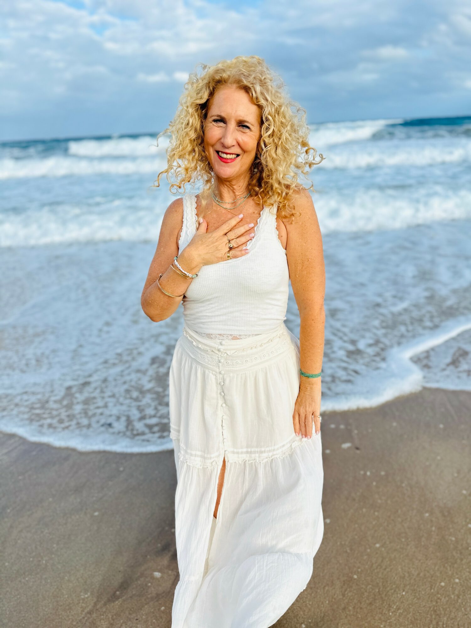 Woman with curly blonde hair smiling on the beach, wearing a white dress, with ocean waves in background.