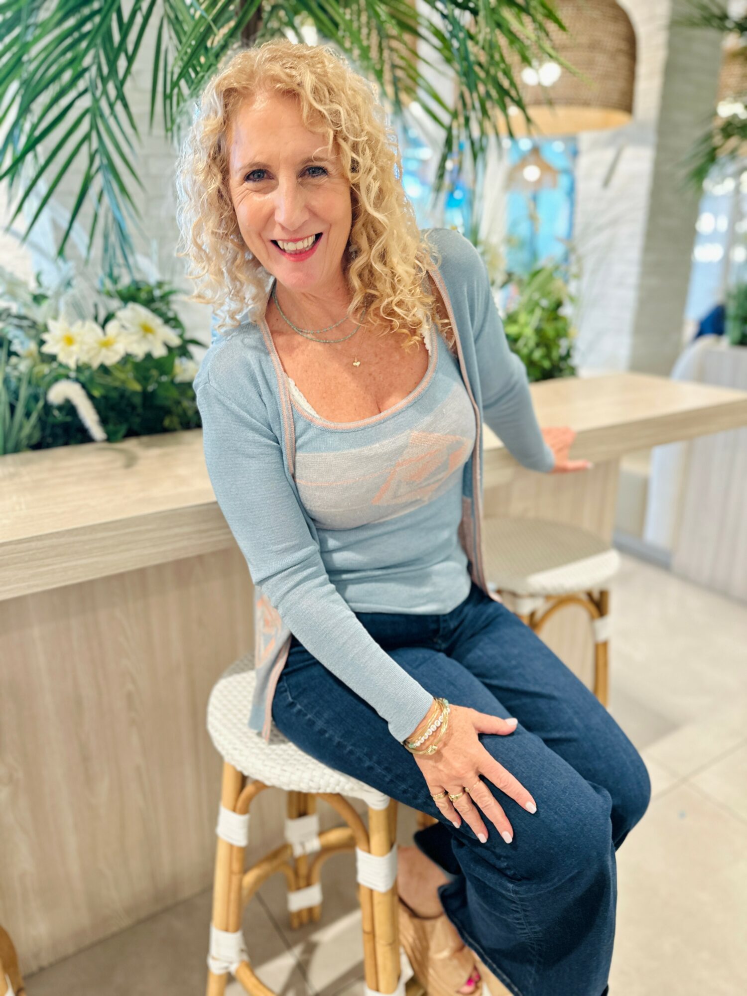 Woman with curly blonde hair sitting on a stool in a bright indoor space, smiling, with plants and flowers behind her.