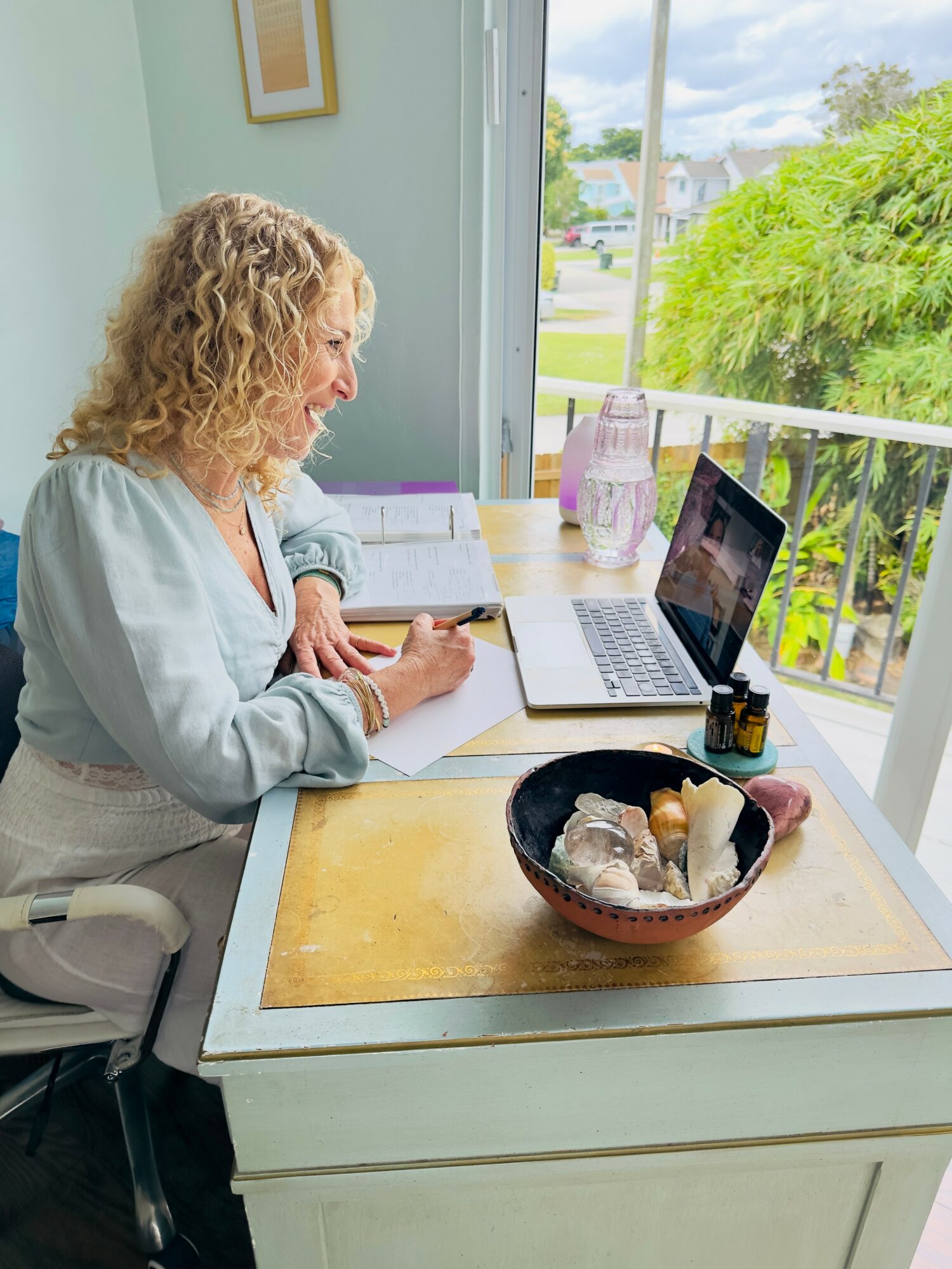 Woman with curly hair working at a desk with a laptop, notebook, and a bowl of shells, near a window with greenery outside.