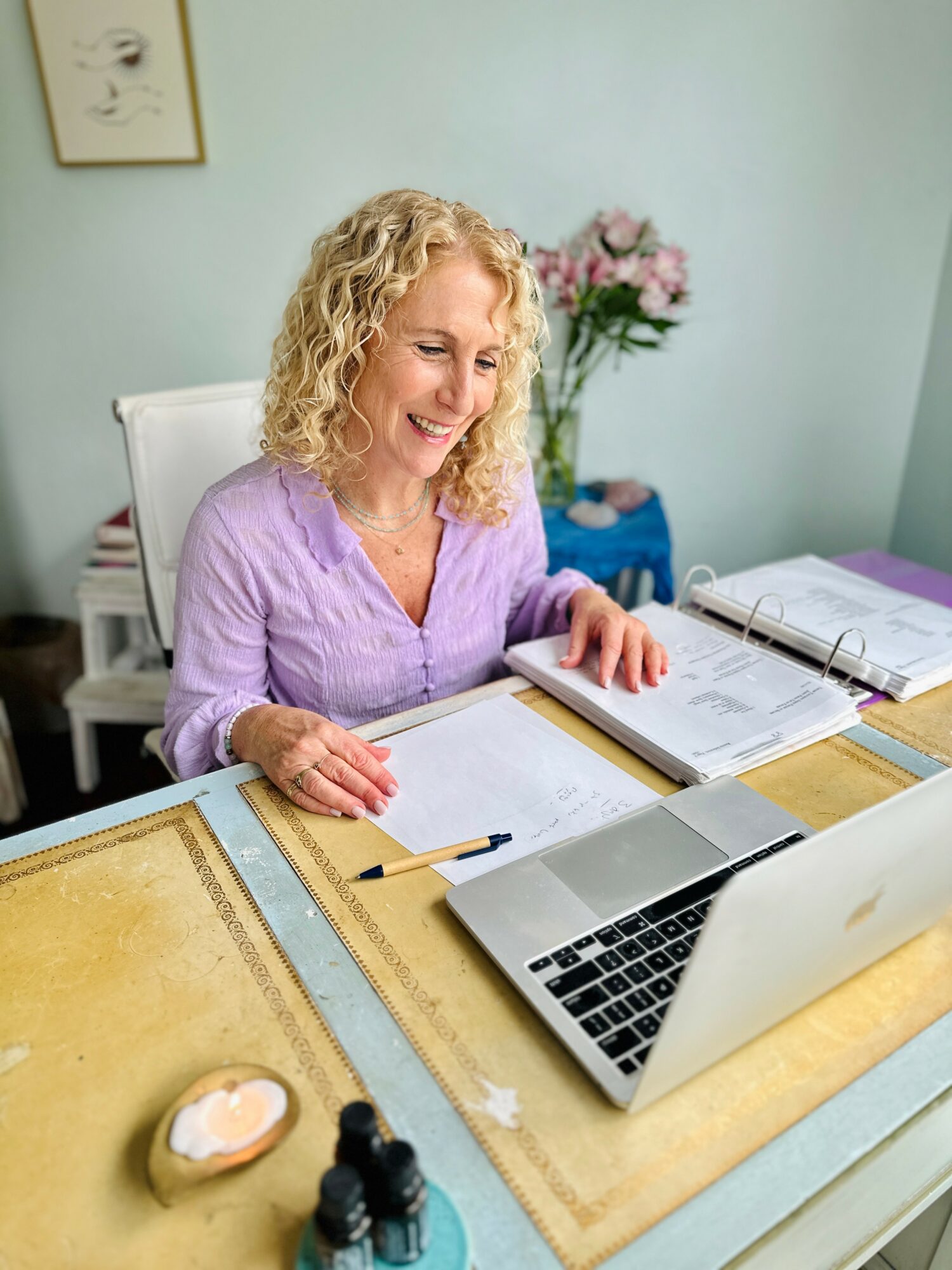 Woman with curly blonde hair smiling at a laptop with open notebooks on a desk, in a room with a flower vase and framed art.