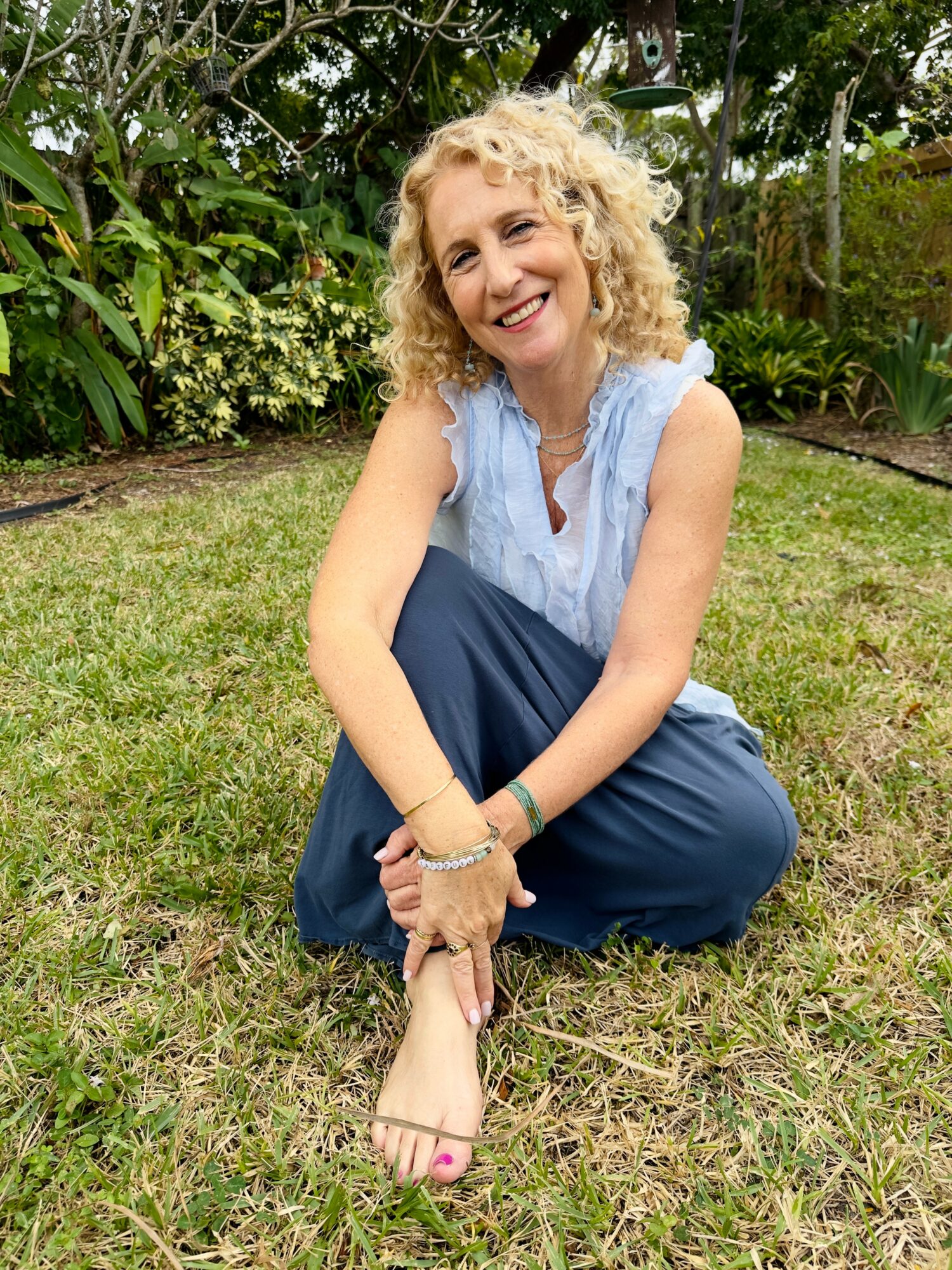 Woman with curly blonde hair sitting on grass in garden, smiling, wearing sleeveless light blue top and dark blue pants.