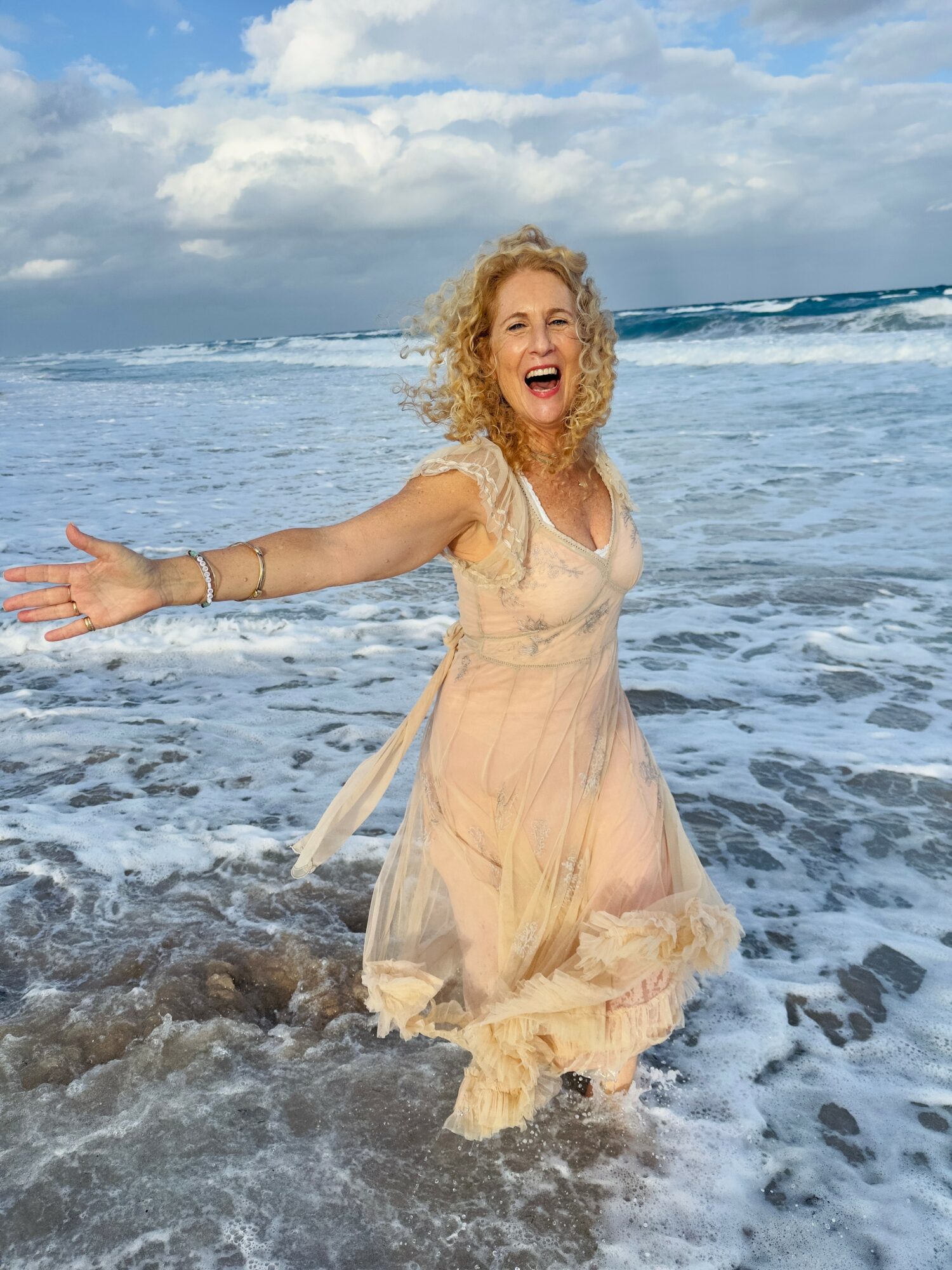 Woman with curly hair in a light dress standing in ocean water, smiling with arms outstretched.