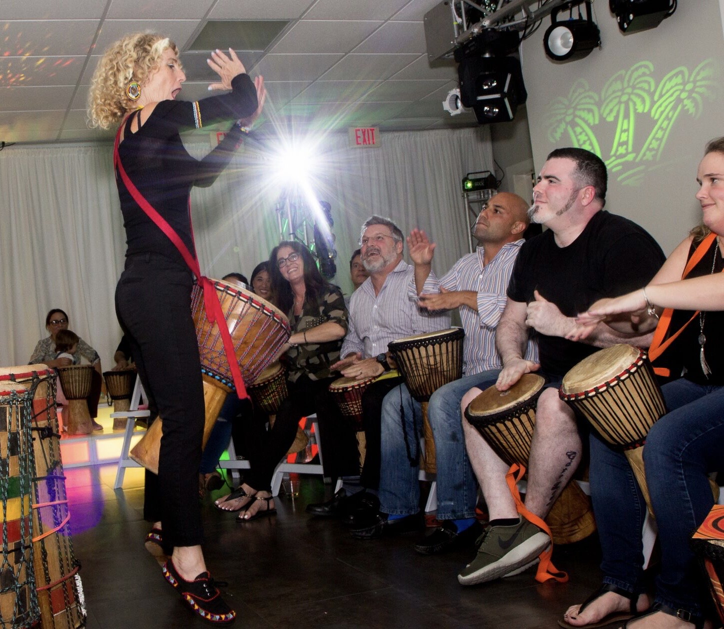 Woman leading a drumming circle with people playing drums in a room with colorful lighting.