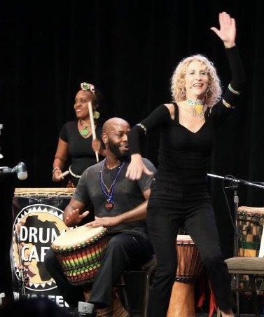 Three musicians performing on stage, one woman singing with hand raised, two others playing drums, black background.