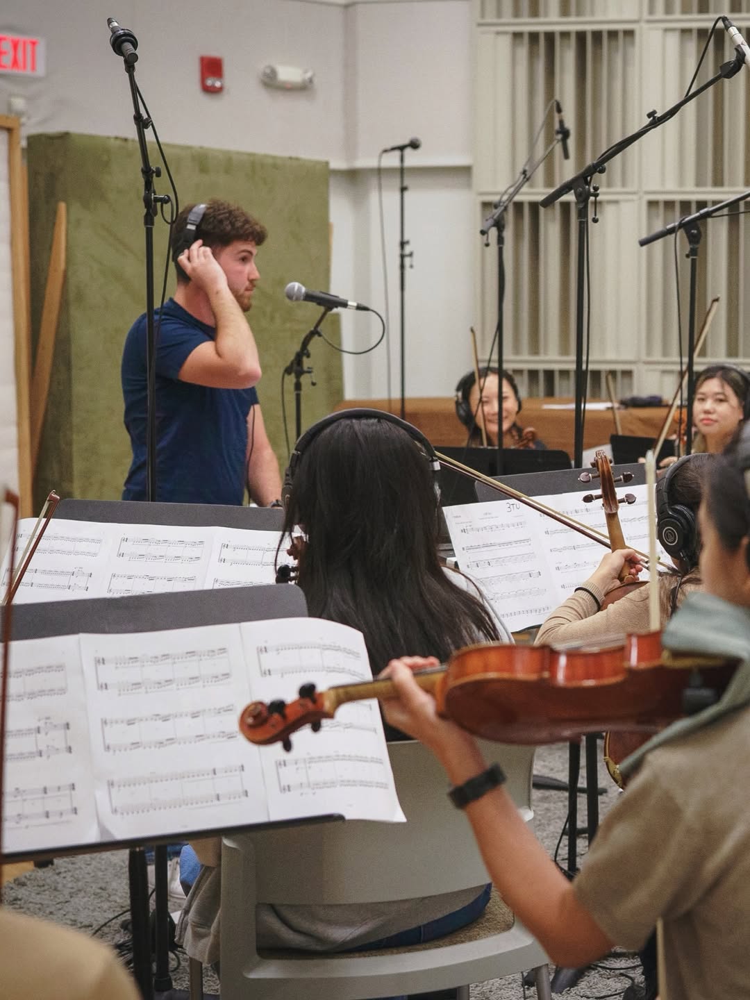 Young man with headphones standing near music stands and orchestra members in a rehearsal room.