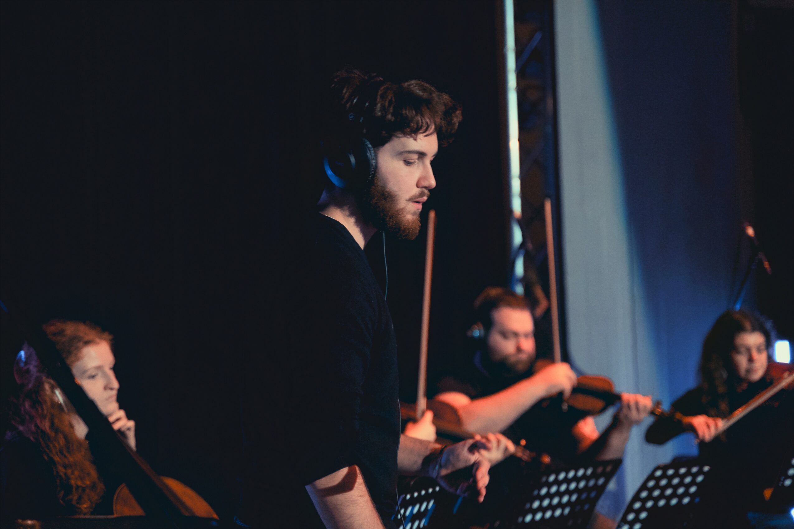 Young man with headphones playing violin on stage with other musicians in background.
