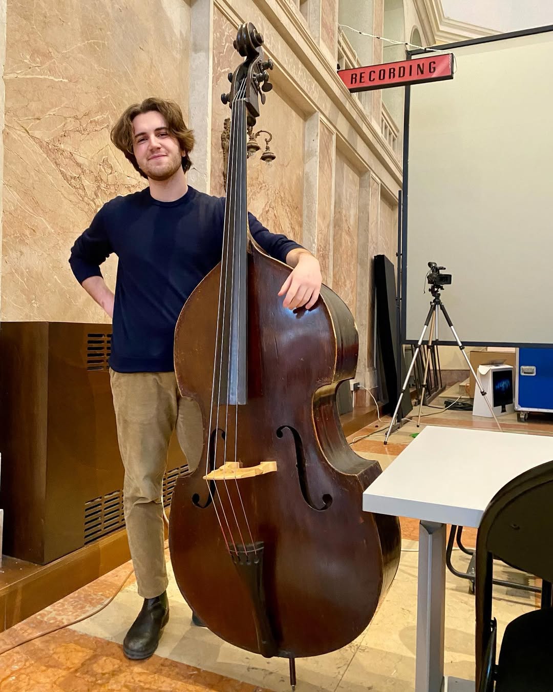 Young man standing next to a large double bass in a recording studio, with recording equipment and a tripod in the background.