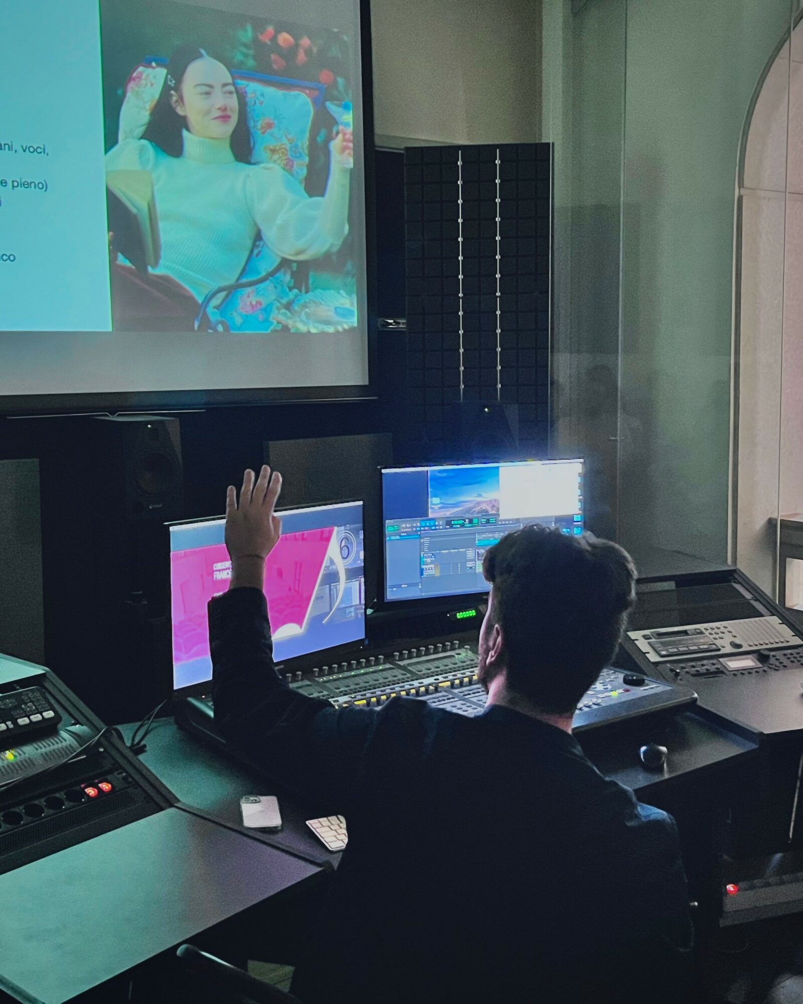 Person with dark hair and dark clothing raising hand in front of multiple computer screens in a control room.