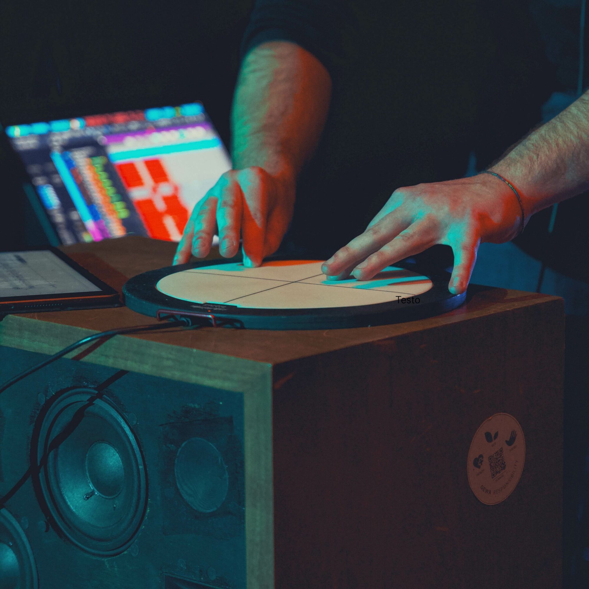 Person's hands playing a drum pad on a wooden table with audio equipment and a laptop in background.