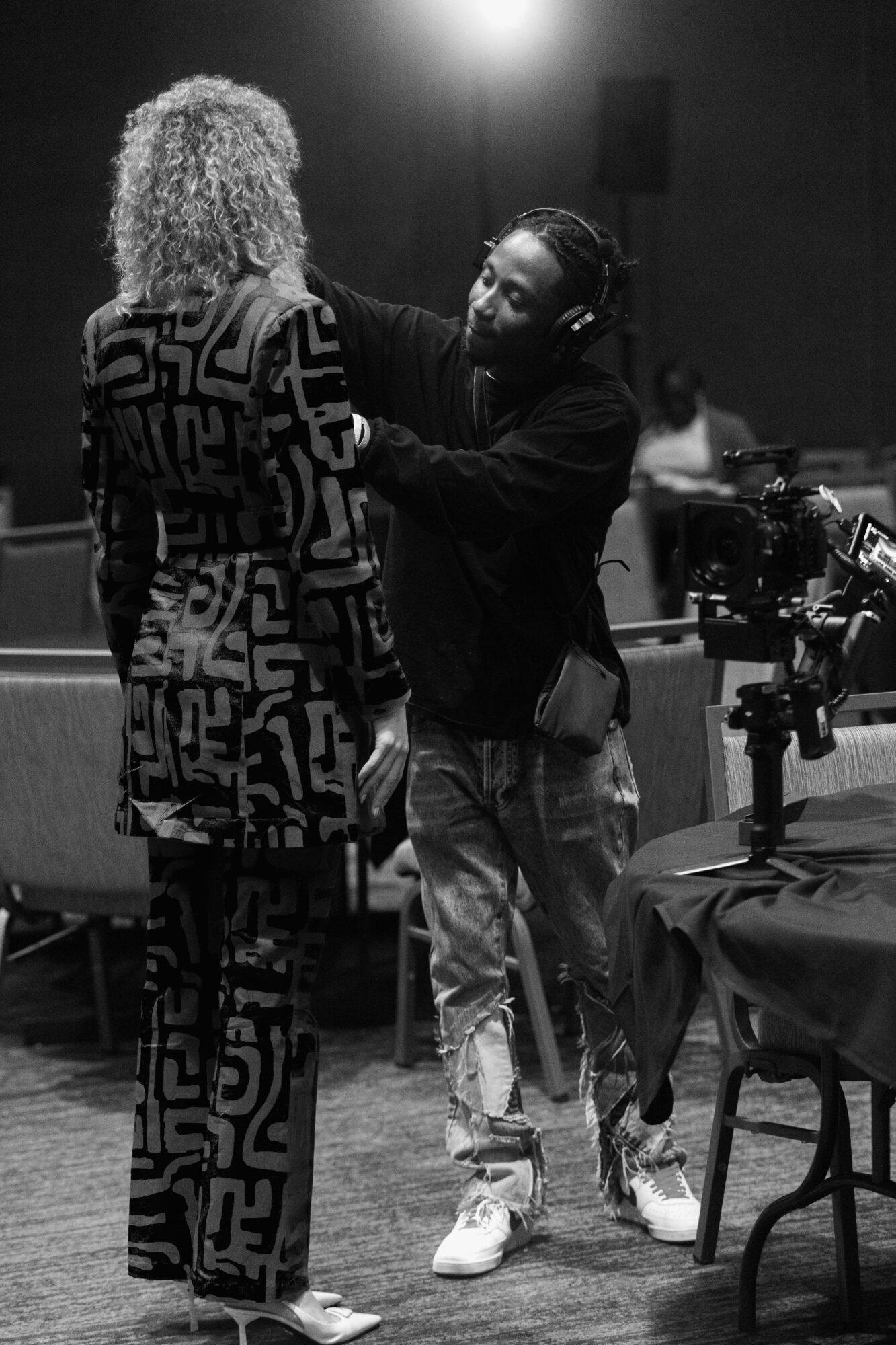 Two people in conversation, one adjusting the other's clothing, in a room with a camera and equipment.
