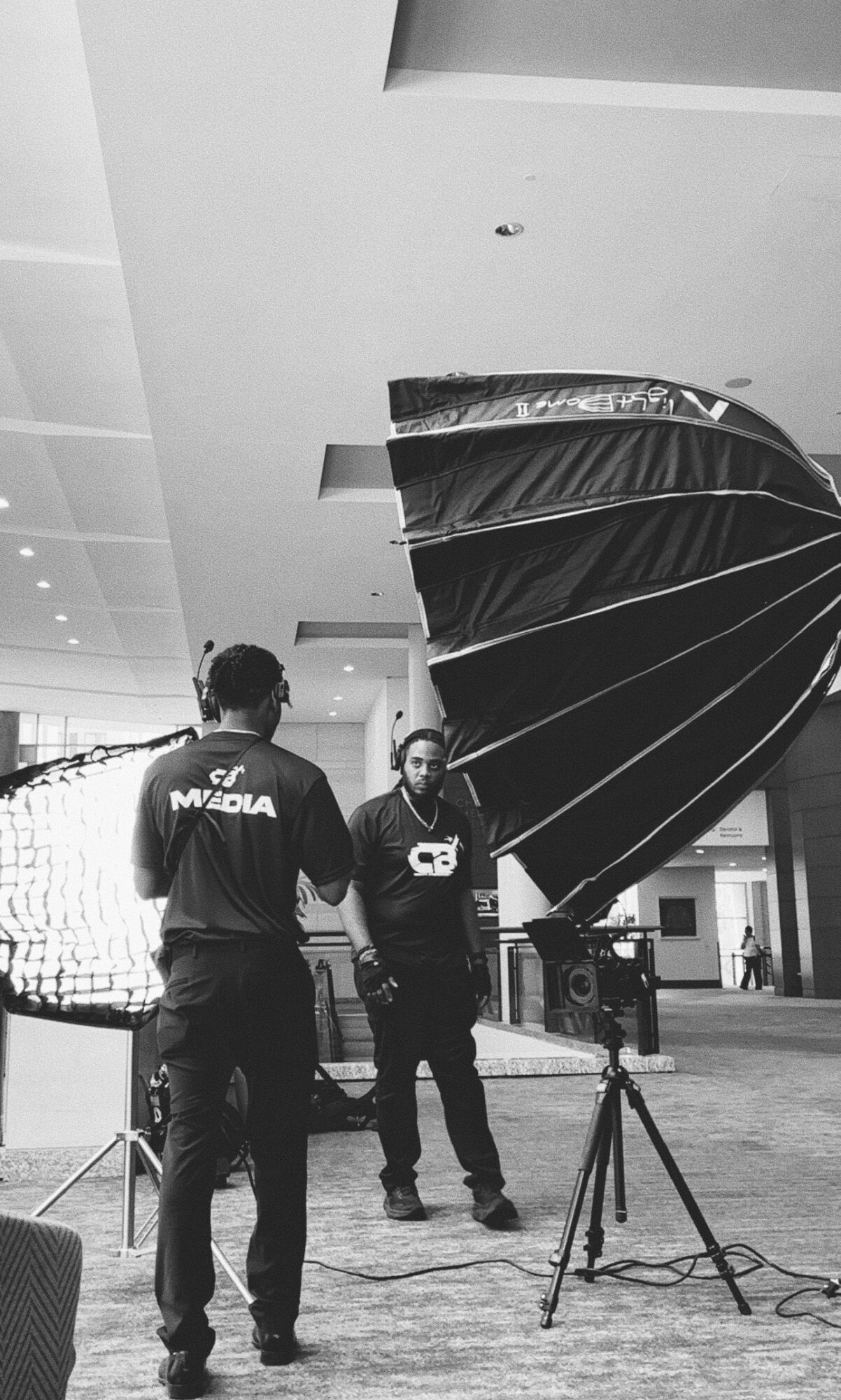 Two people in a spacious indoor area with a large black umbrella and photography equipment.