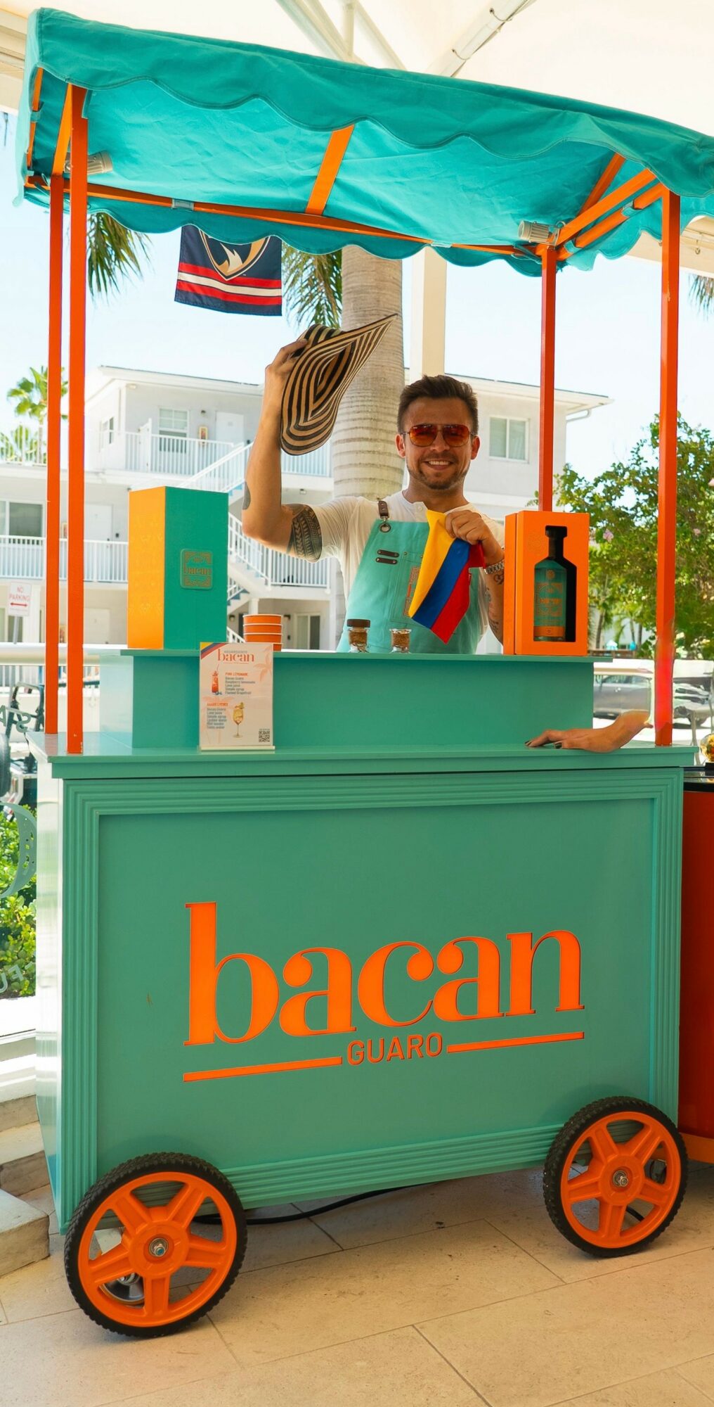 Person standing behind a teal Bacardi-branded cart with orange wheels, waving and smiling outdoors.