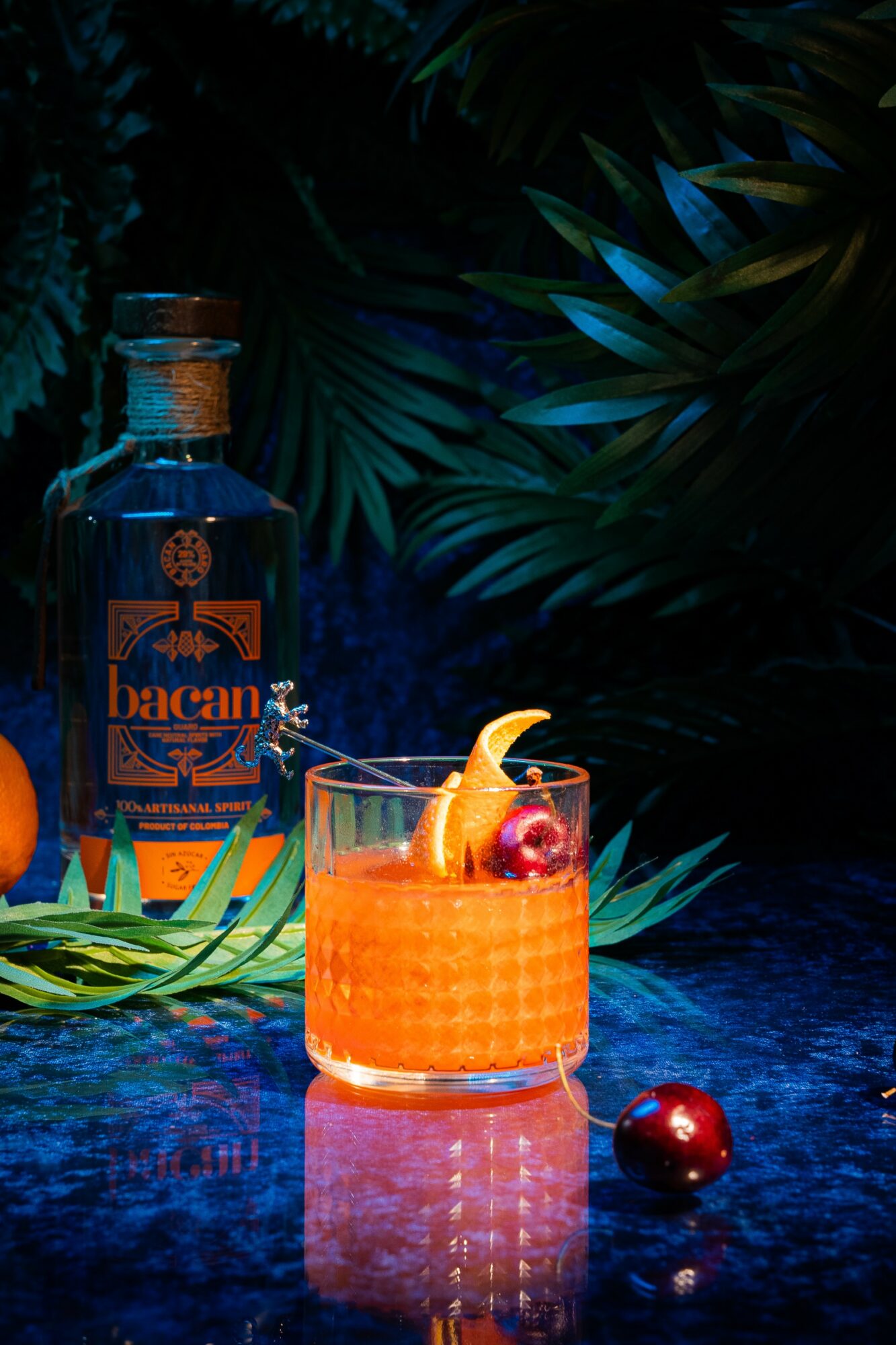 A glass with orange-colored drink garnished with lemon peel and cherries, placed on a reflective surface with a Bacardi bottle in background.