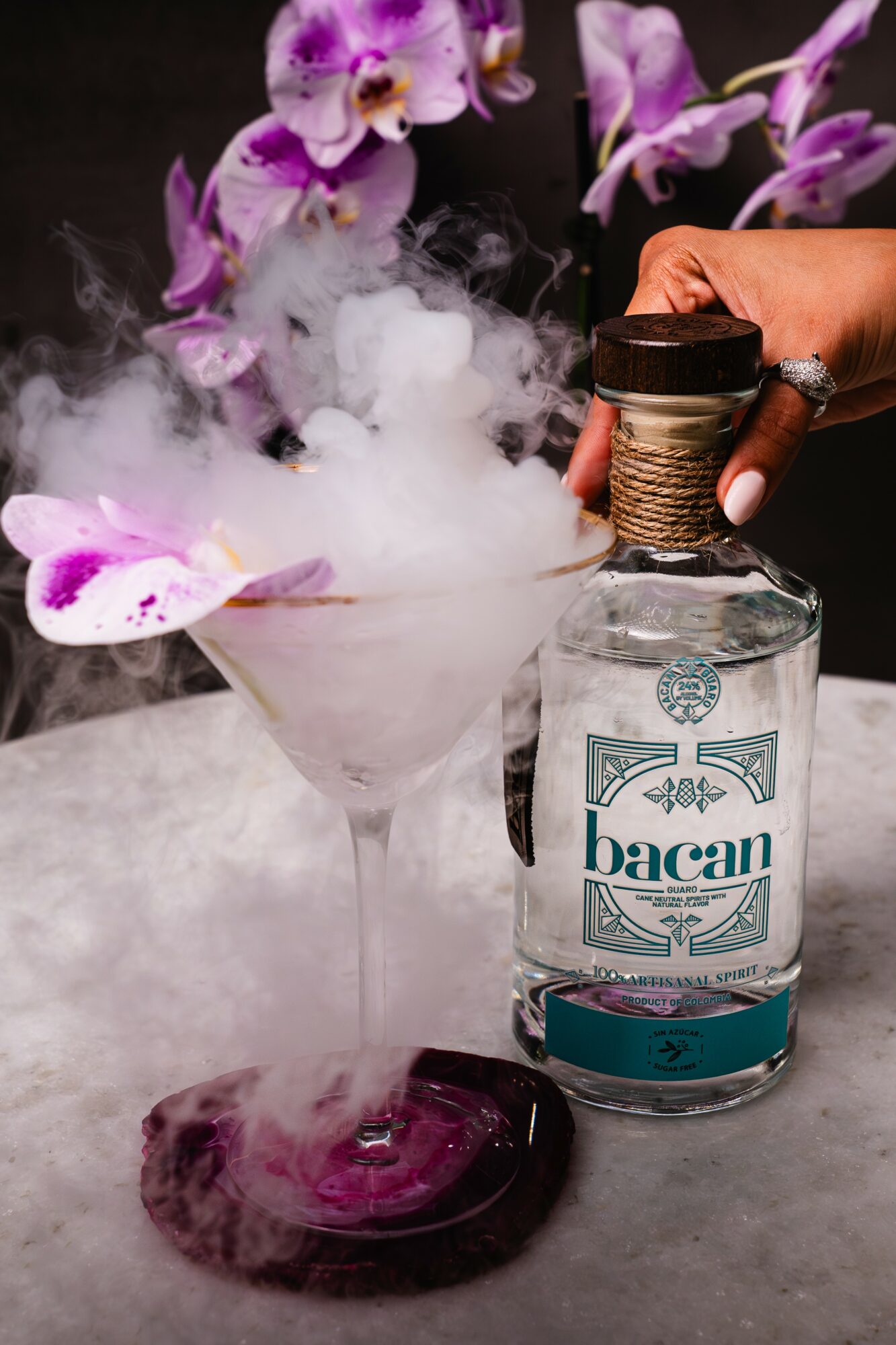Hand holding Bacardi bottle near a cocktail glass with purple orchids and dry ice smoke.