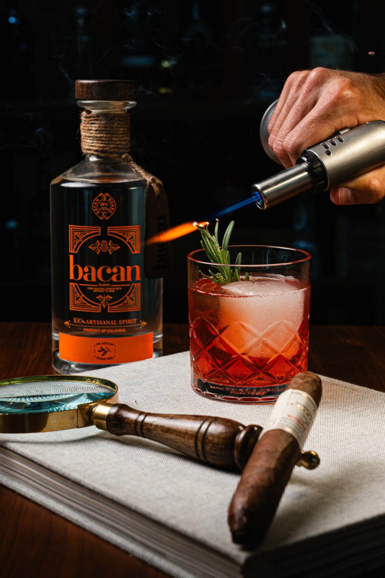 Person using a torch to garnish a pink cocktail with herbs, on a table with a bottle and tools.