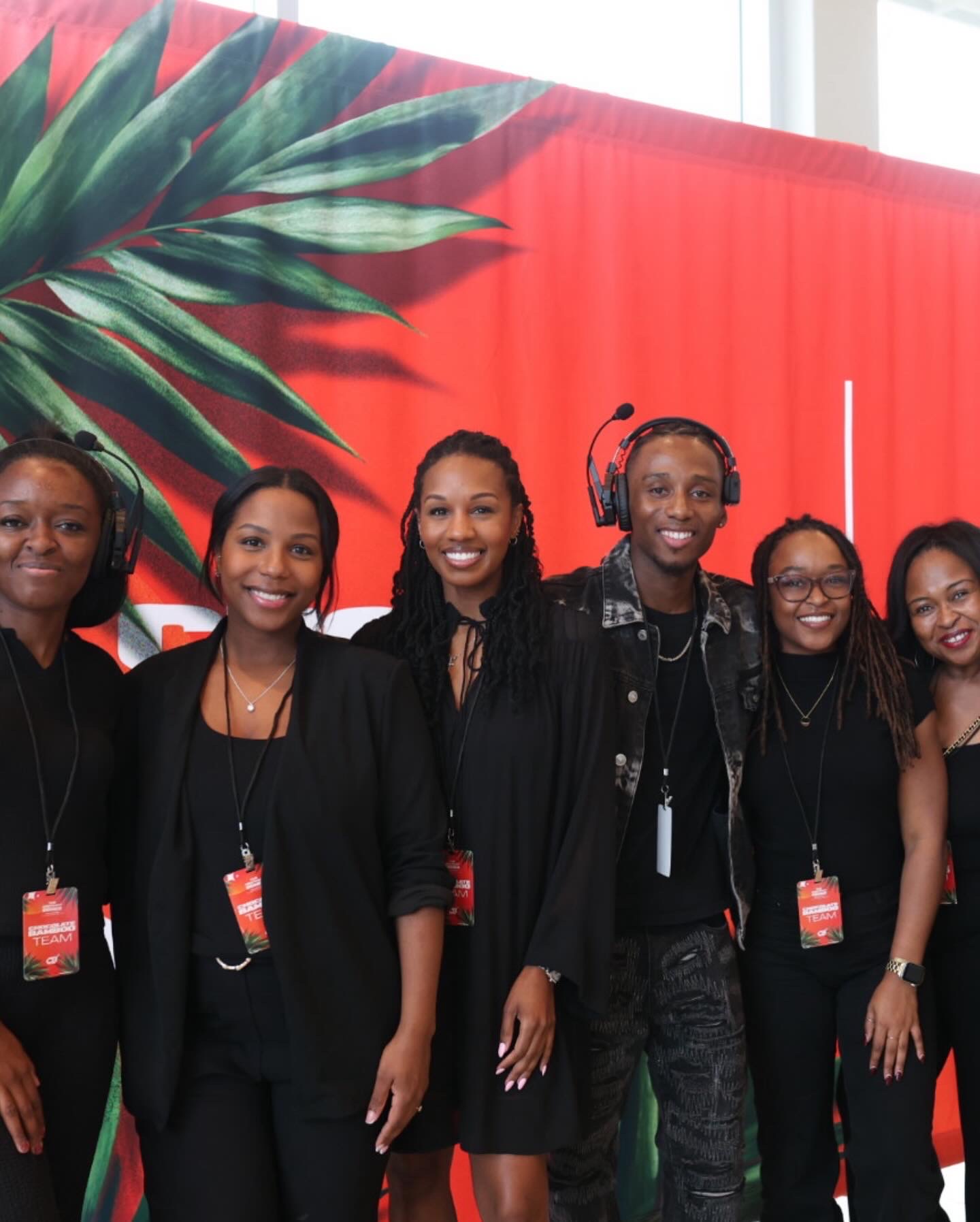 Group of six women standing together in front of a red and green tropical-themed backdrop, smiling at the camera.